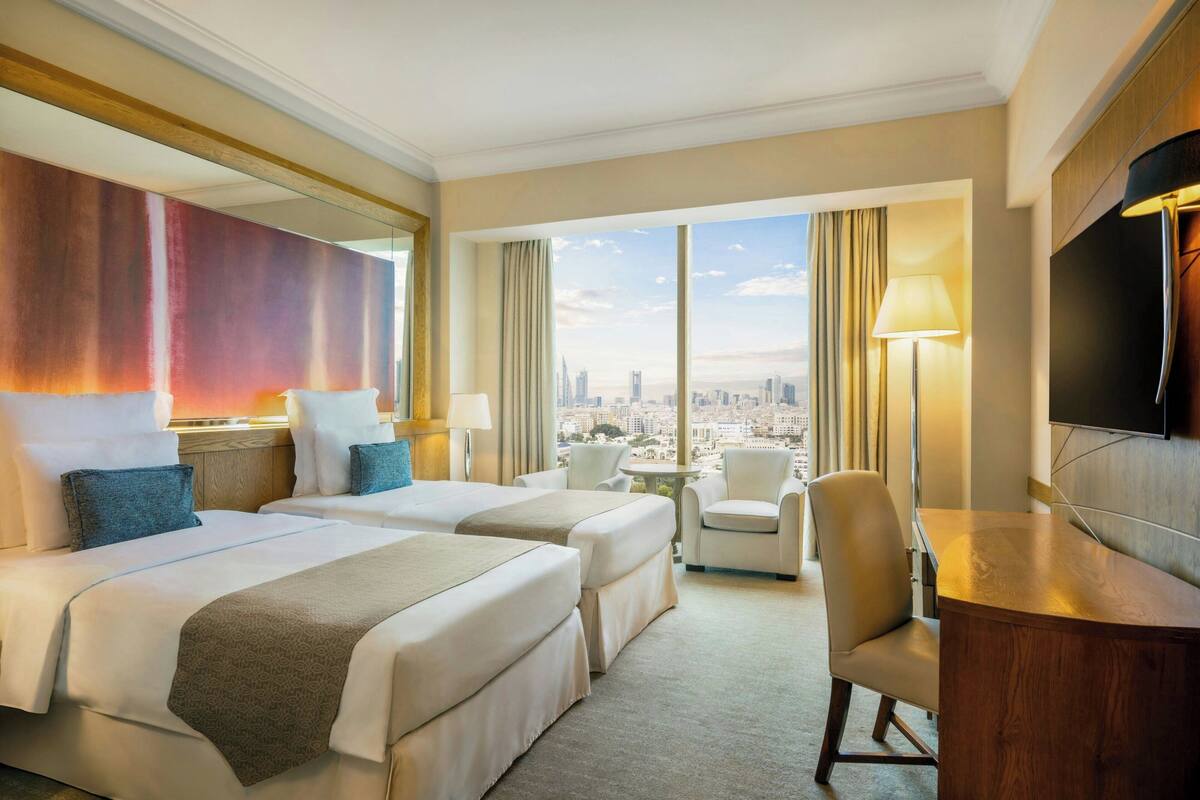 A hotel room with two beds, a desk, a chair, a TV, and a view of the city.