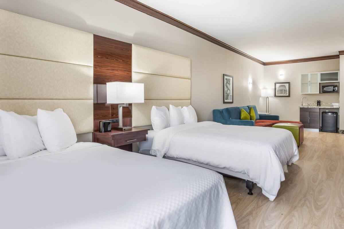 A hotel room with two beds, a sofa, a desk, a lamp, a microwave, and a small kitchen area.