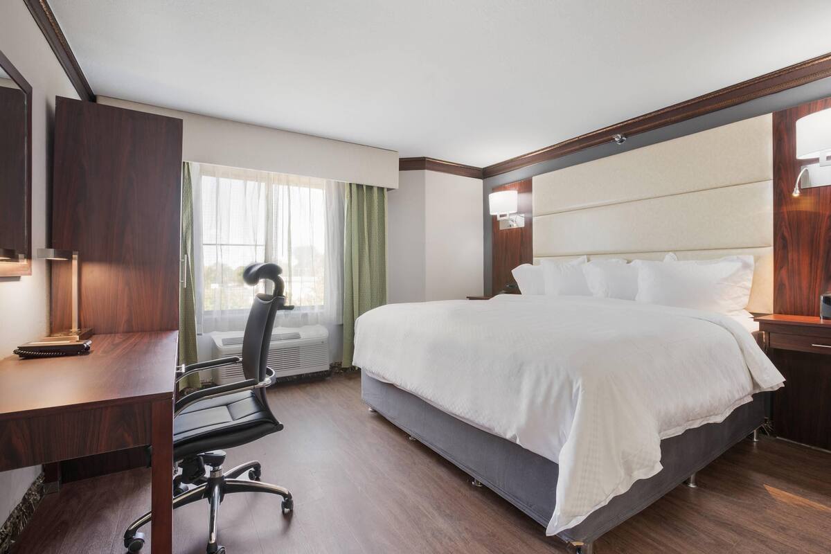 A hotel room with a large bed, a desk with a chair, a window with curtains, and a nightstand with a lamp.