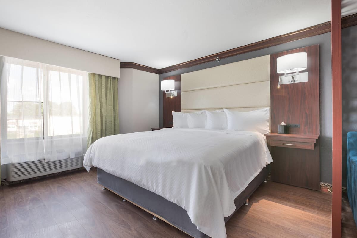 A hotel room with a large bed, a bedside table with a lamp, a window with sheer curtains, and a wooden floor.