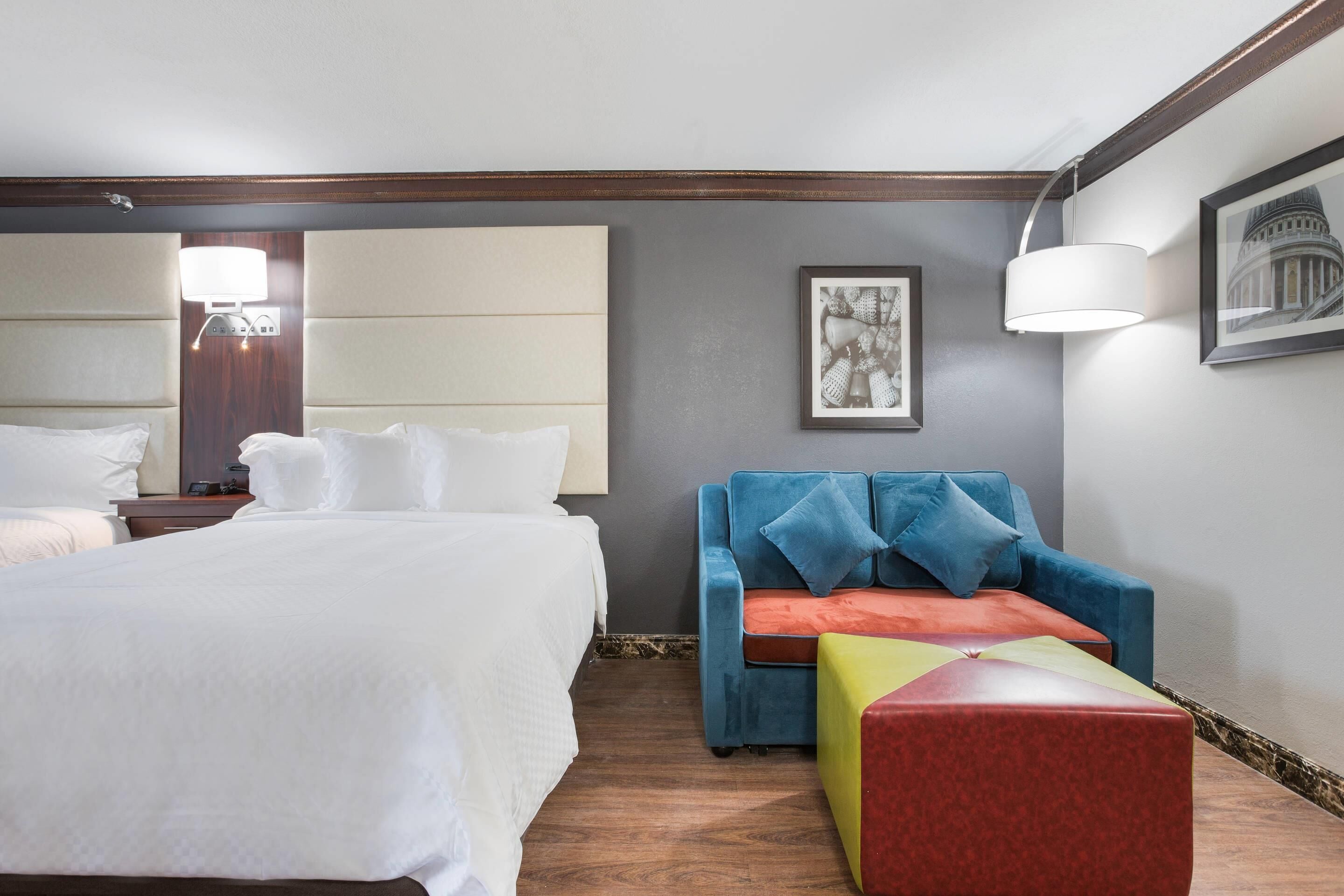 A hotel room with a large bed, a blue sofa with a red and yellow ottoman, a framed picture on the wall, and a lamp.