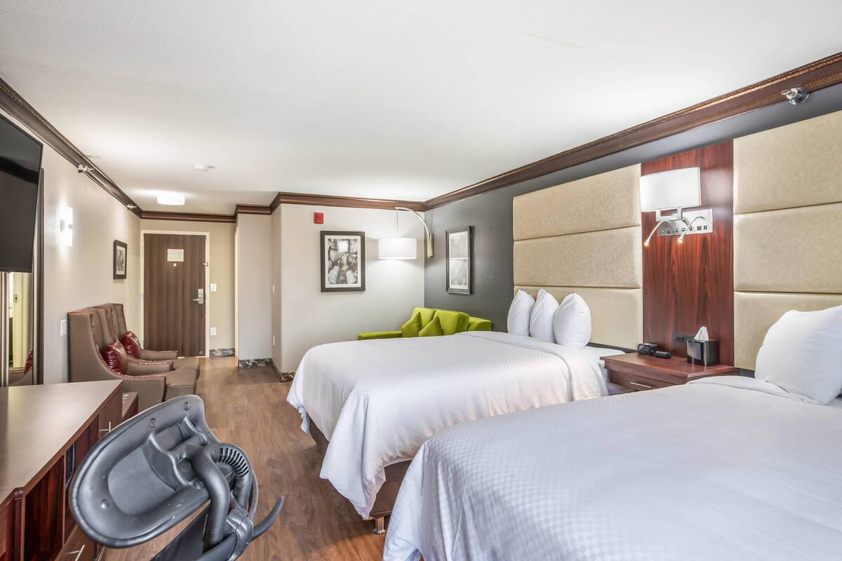 A hotel room with two beds, a desk, a chair, and a small seating area with a green chair.