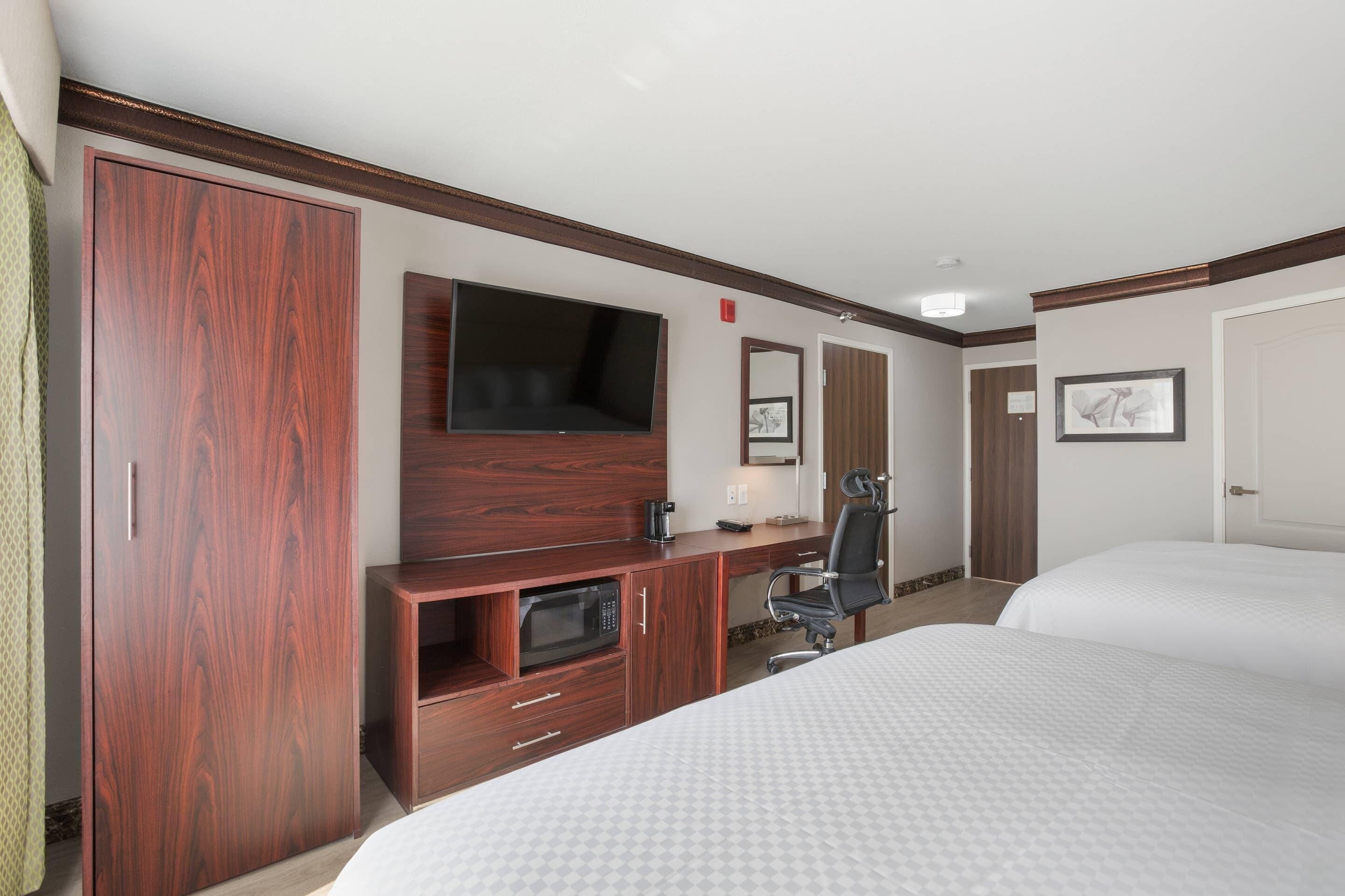 A hotel room with a large wardrobe, a flat-screen TV, a microwave, a desk with a chair, and two beds.