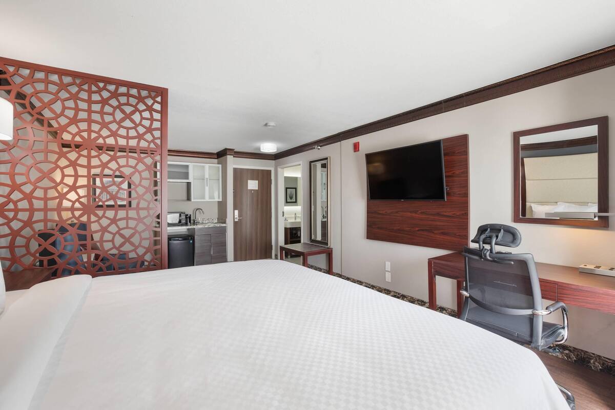 A hotel room with a large bed, a desk with a chair, a TV mounted on the wall, and a bathroom visible through an open door.