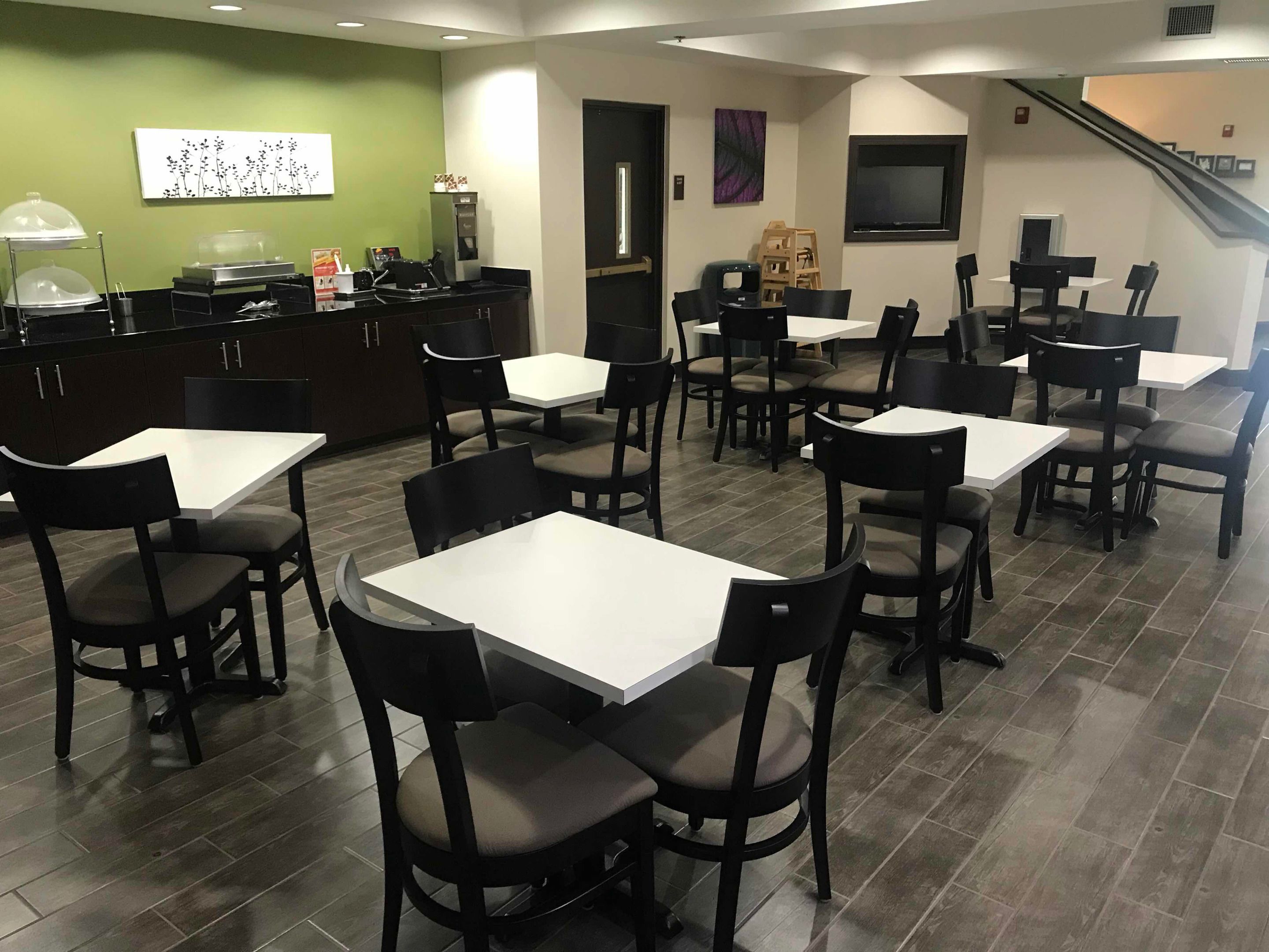 A restaurant with multiple tables and chairs, a counter with food service equipment, and a staircase in the background.