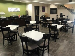 A restaurant with multiple tables and chairs, a counter with food service equipment, and a staircase in the background.
