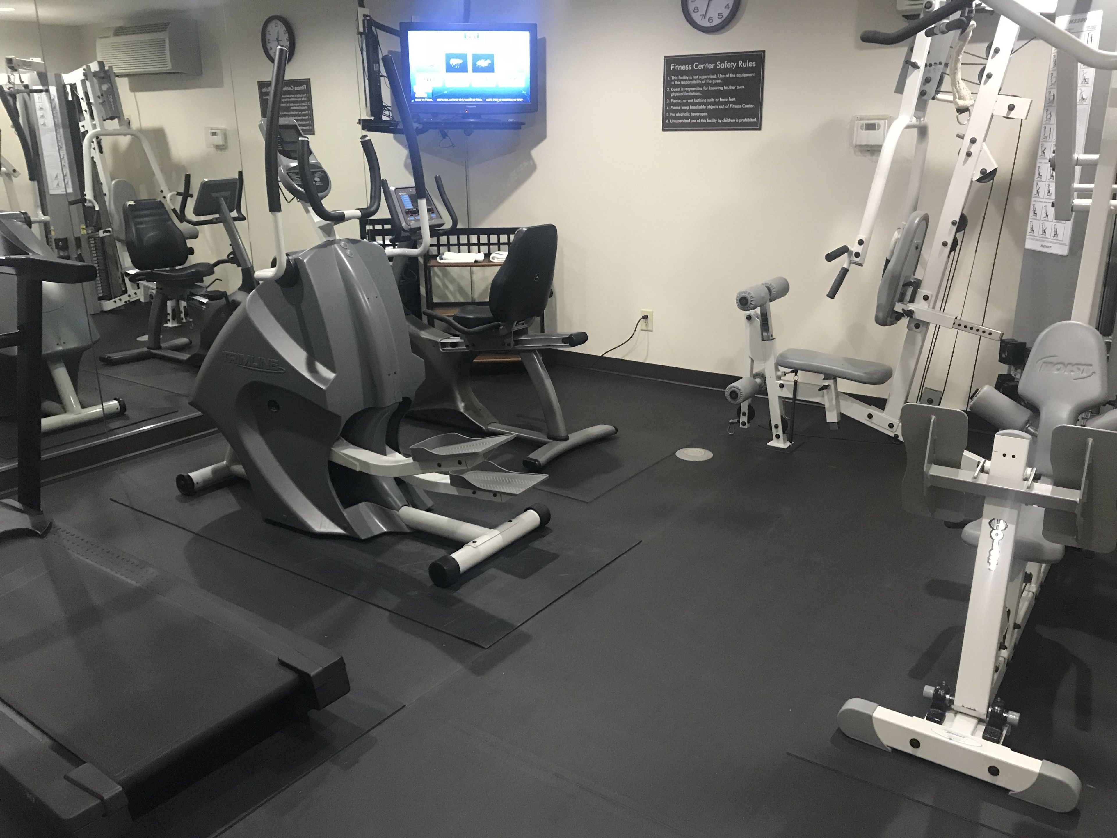 Ein Fitnessraum mit unterschiedlichen Trainingsgeräten, einem Fernsehbildschirm und einem Hinweis zu den Sicherheitsbestimmungen an der Wand.