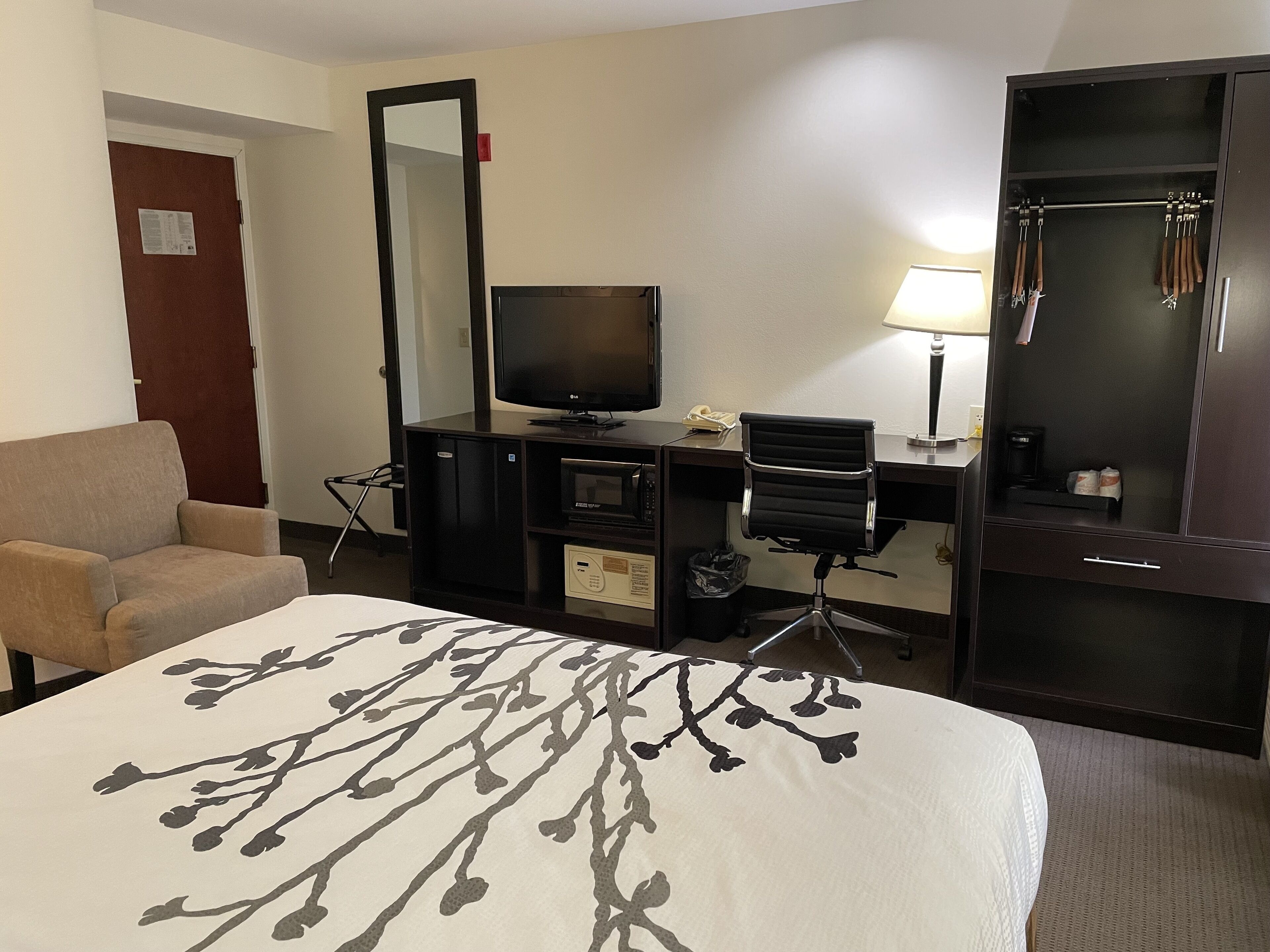 A hotel room with a bed, a desk with a computer, a TV, a chair, a coat rack, and a mirror.