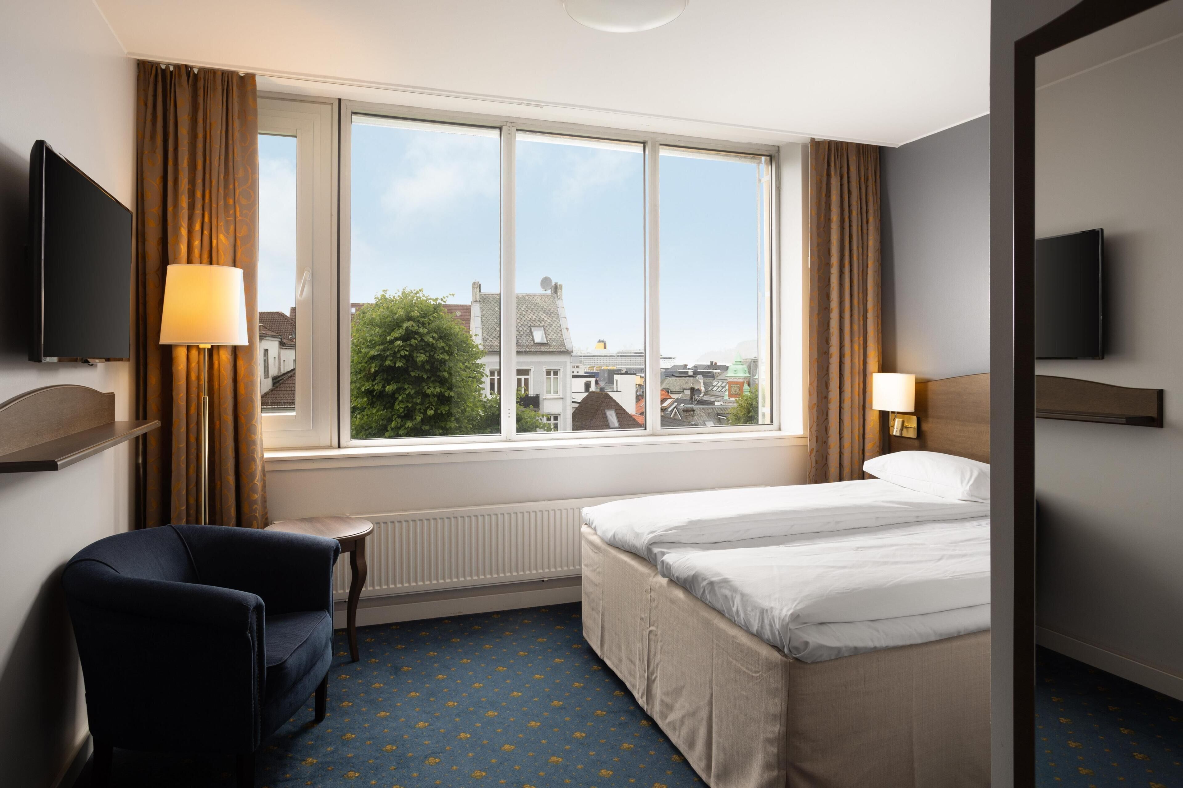Habitación de hotel con cama, una silla, televisión, ventana con cortinas y vistas a edificios.