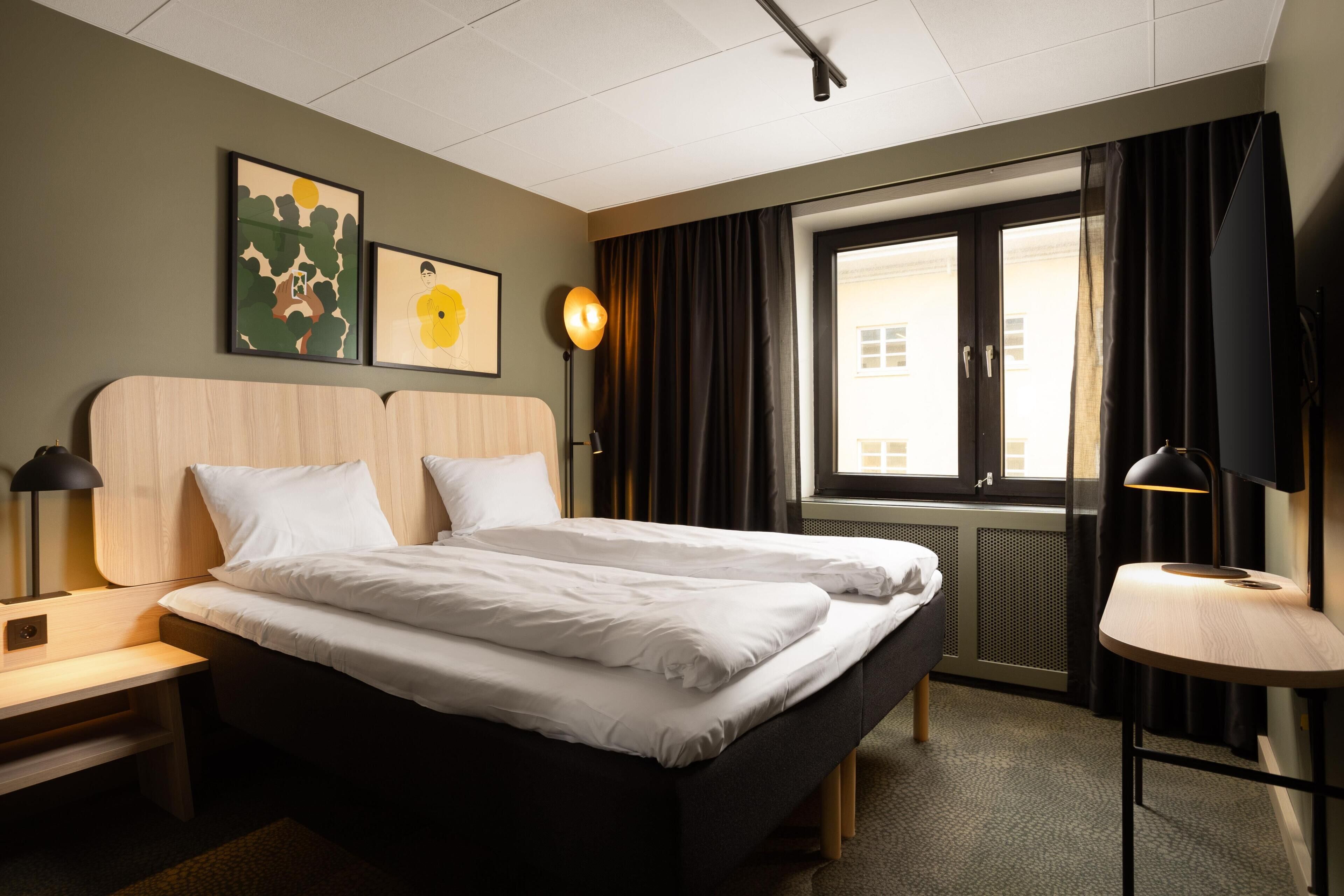A hotel room with two beds, a desk, a lamp, a TV, and artwork on the wall.