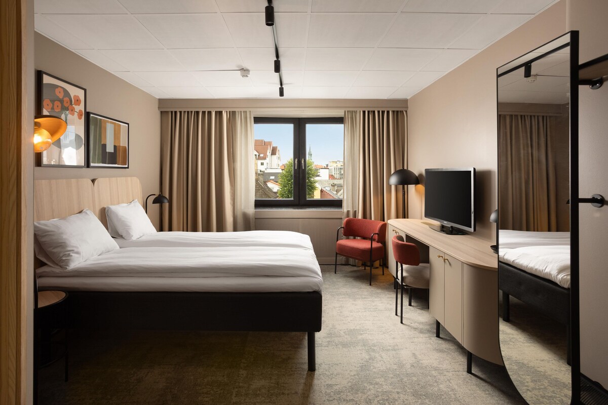 Habitación de hotel con dos camas, un escritorio con televisor, una silla y una ventana con cortinas.