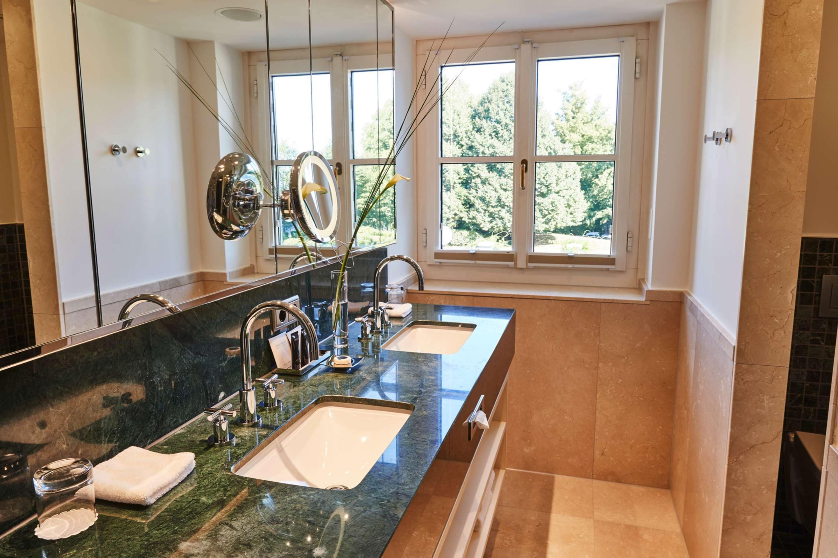 Ein modernes Badezimmer mit zwei Waschbecken, einem großen Spiegel und Blick auf Bäume durch die Fenster.