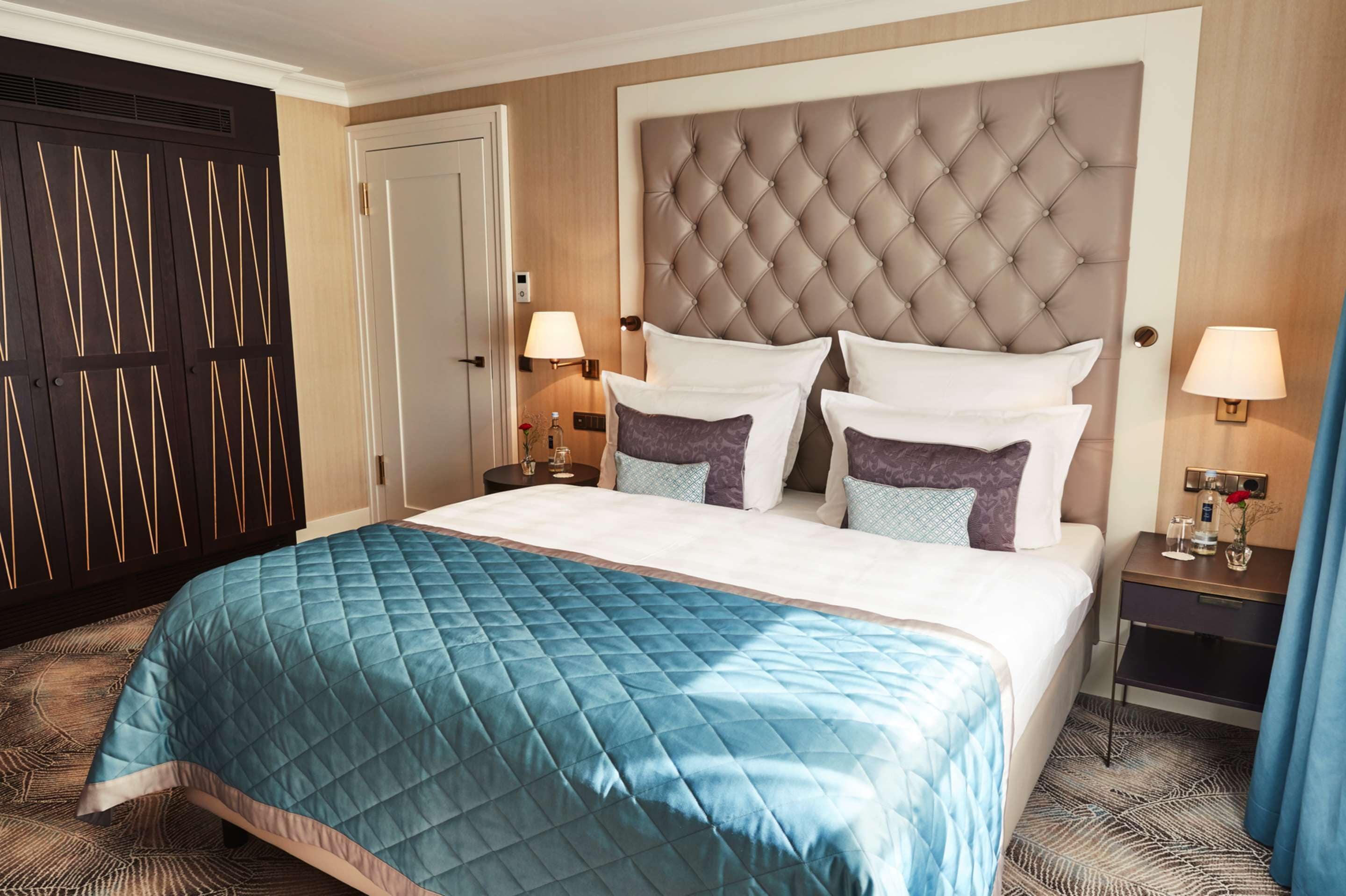 A hotel room with a large bed, a dark wooden wardrobe, a tufted headboard, two bedside tables with lamps, and a patterned carpet.