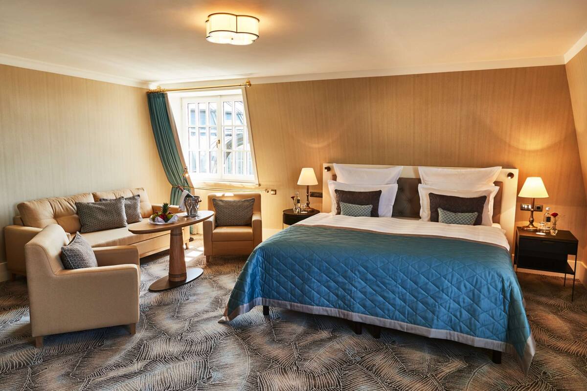 A hotel room with a large bed, a sofa, a chair, a small table, and a window with curtains.
