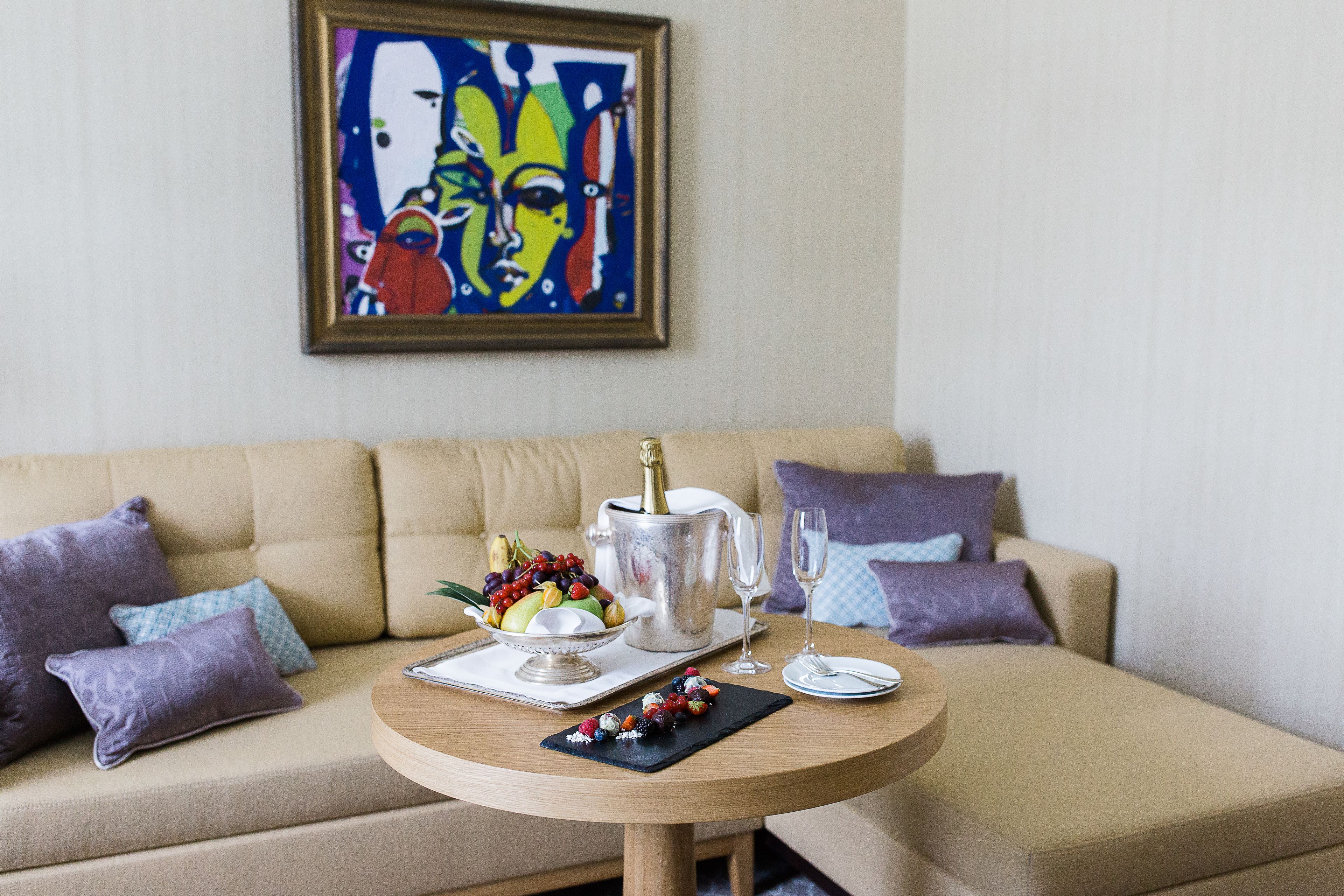 A living room with a beige sofa, a round wooden table with a fruit bowl, a champagne bottle, and two glasses.