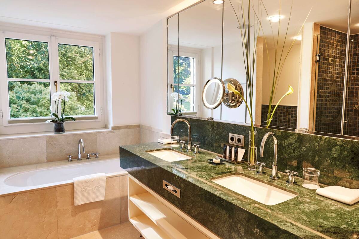 A modern bathroom with two sinks, a large mirror, and a window with a view of greenery.