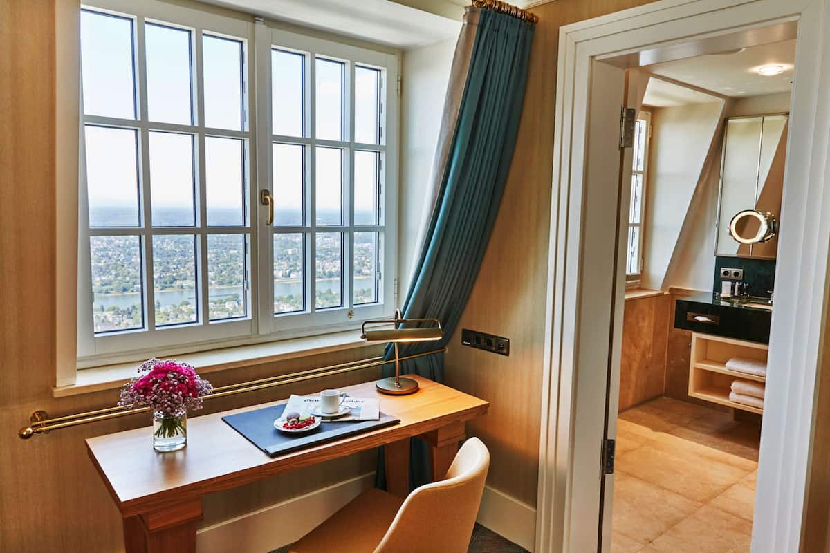 A hotel room with a desk, a chair, a window with a view, a vanity with a mirror, and a bathroom visible through an open door.