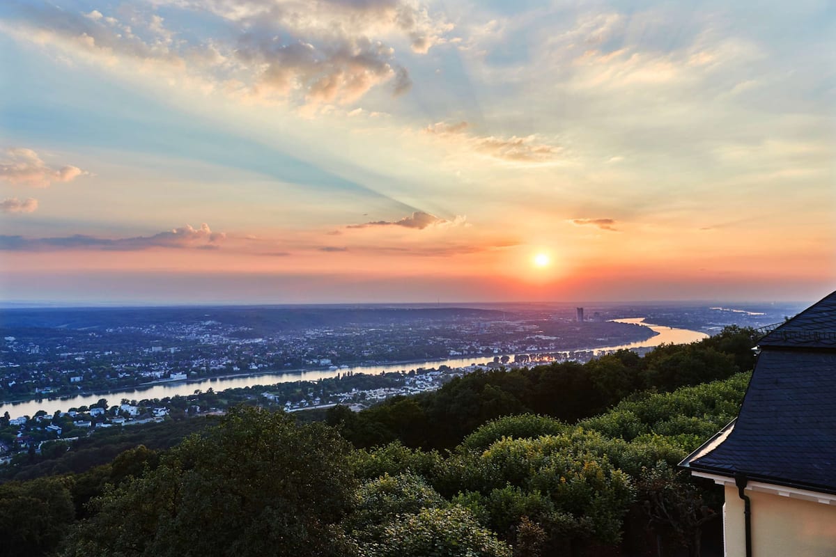 A sunset view over a cityscape with a river, buildings, and trees.