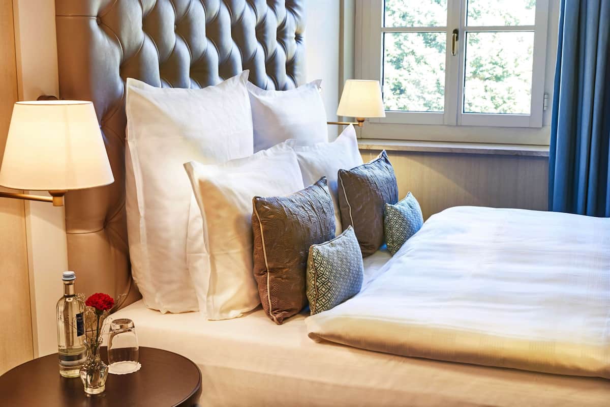 A neatly made bed with a tufted headboard, white and patterned pillows, a bedside table with a lamp, and a window with curtains.