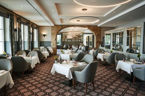 Un comedor espacioso con múltiples mesas y sillas, ventanas amplias y una alfombra estampada.