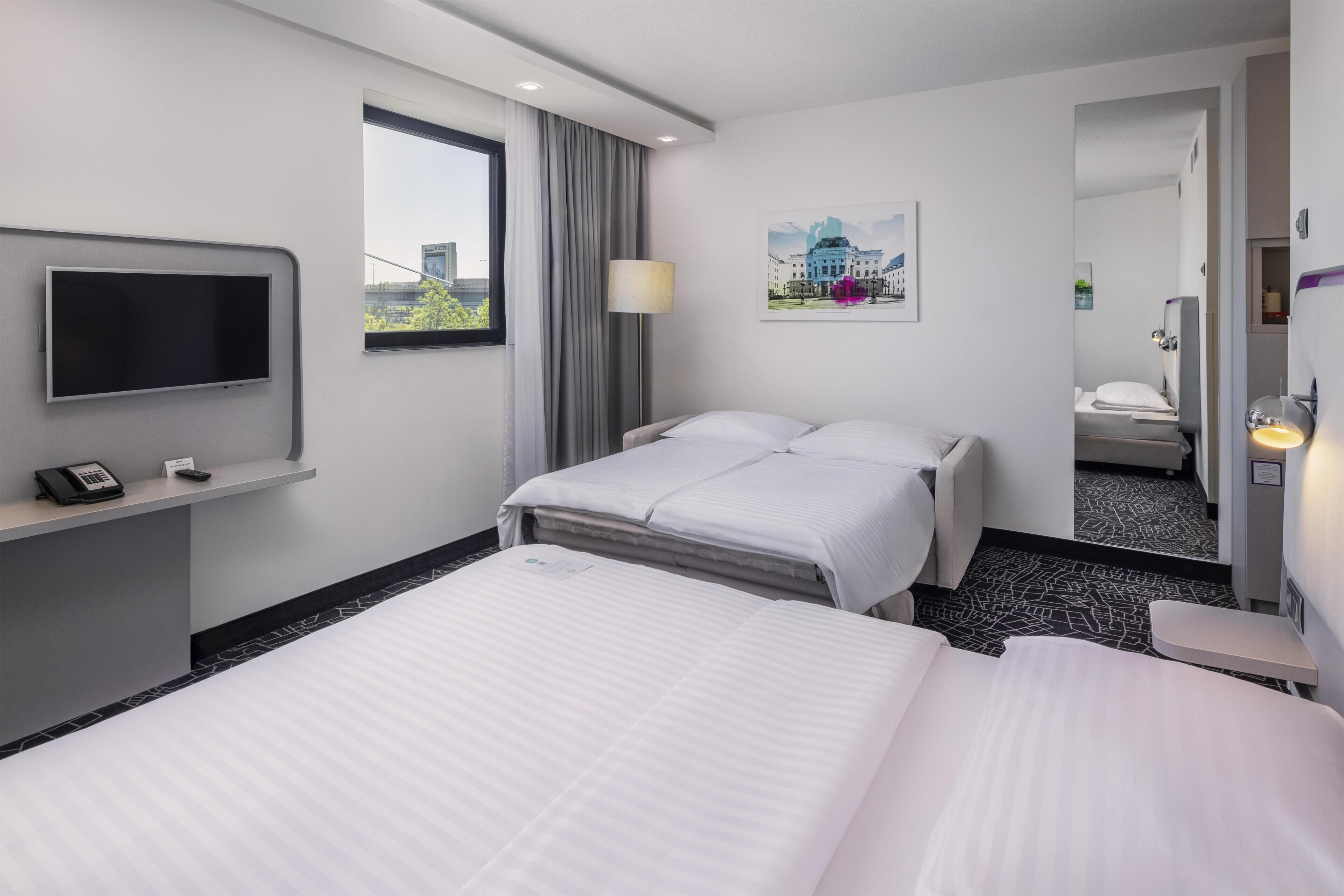 Ein Hotelzimmer mit Bett, Schreibtisch mit Telefon, Fernseher, Fenster mit Vorhängen und einem Bild an der Wand.