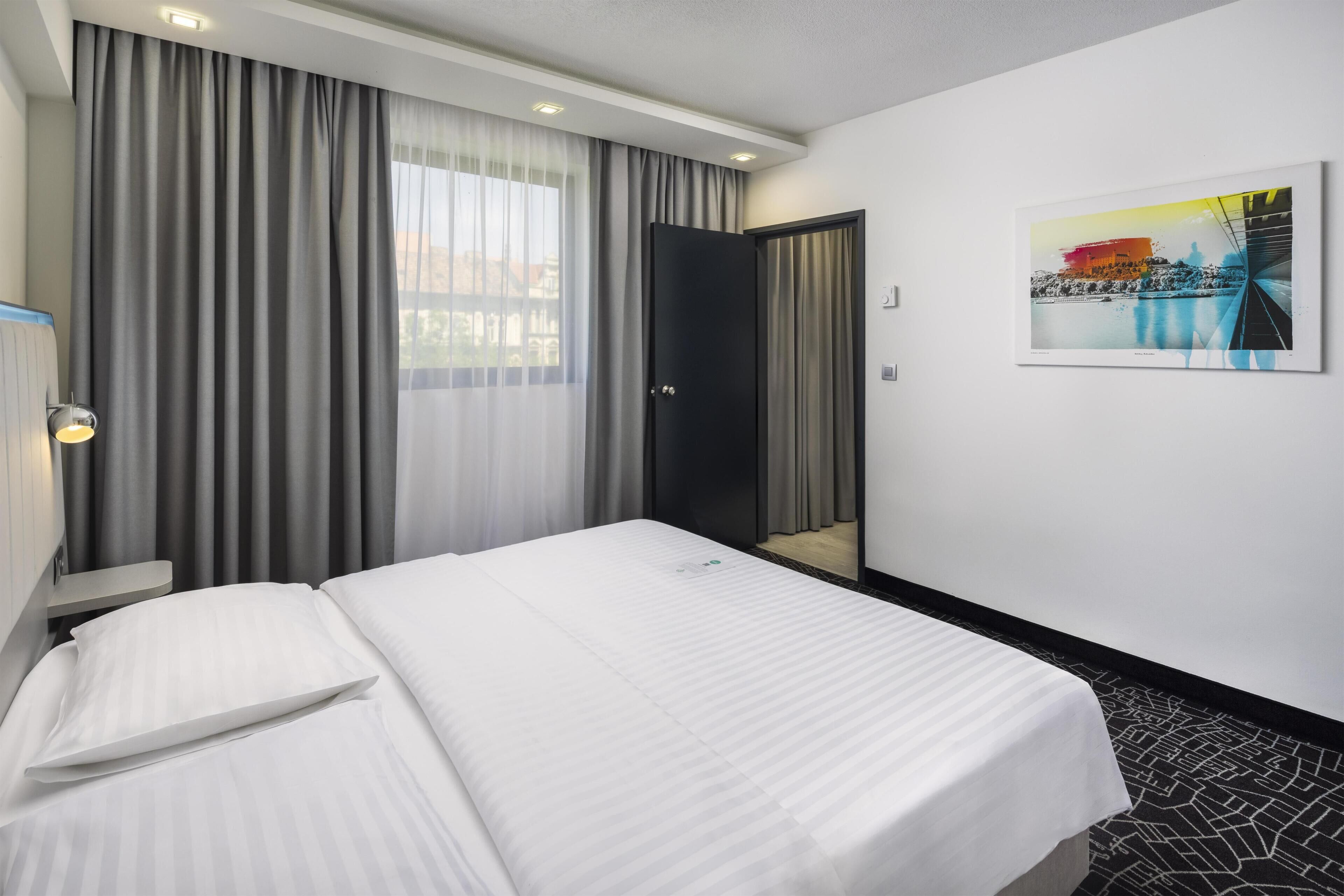 A hotel room with a bed, curtains, a painting, and a door.