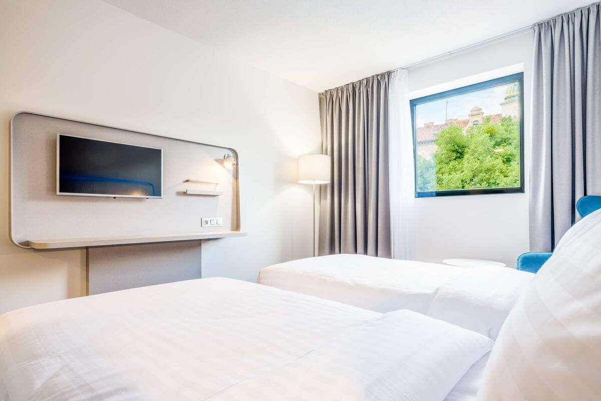 A hotel room with two beds, a flat-screen TV, a lamp, and a window with curtains.