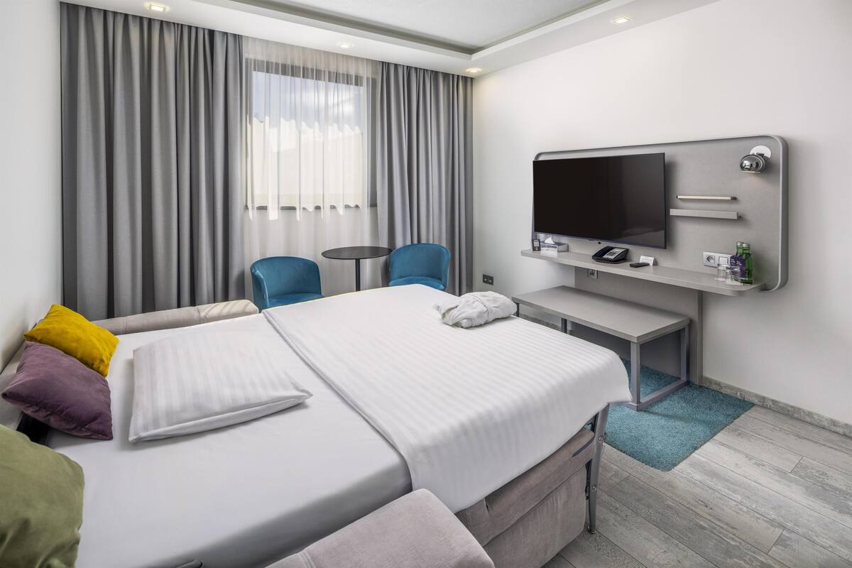 A hotel room with a bed, two chairs, a desk, a TV, and a window with curtains.