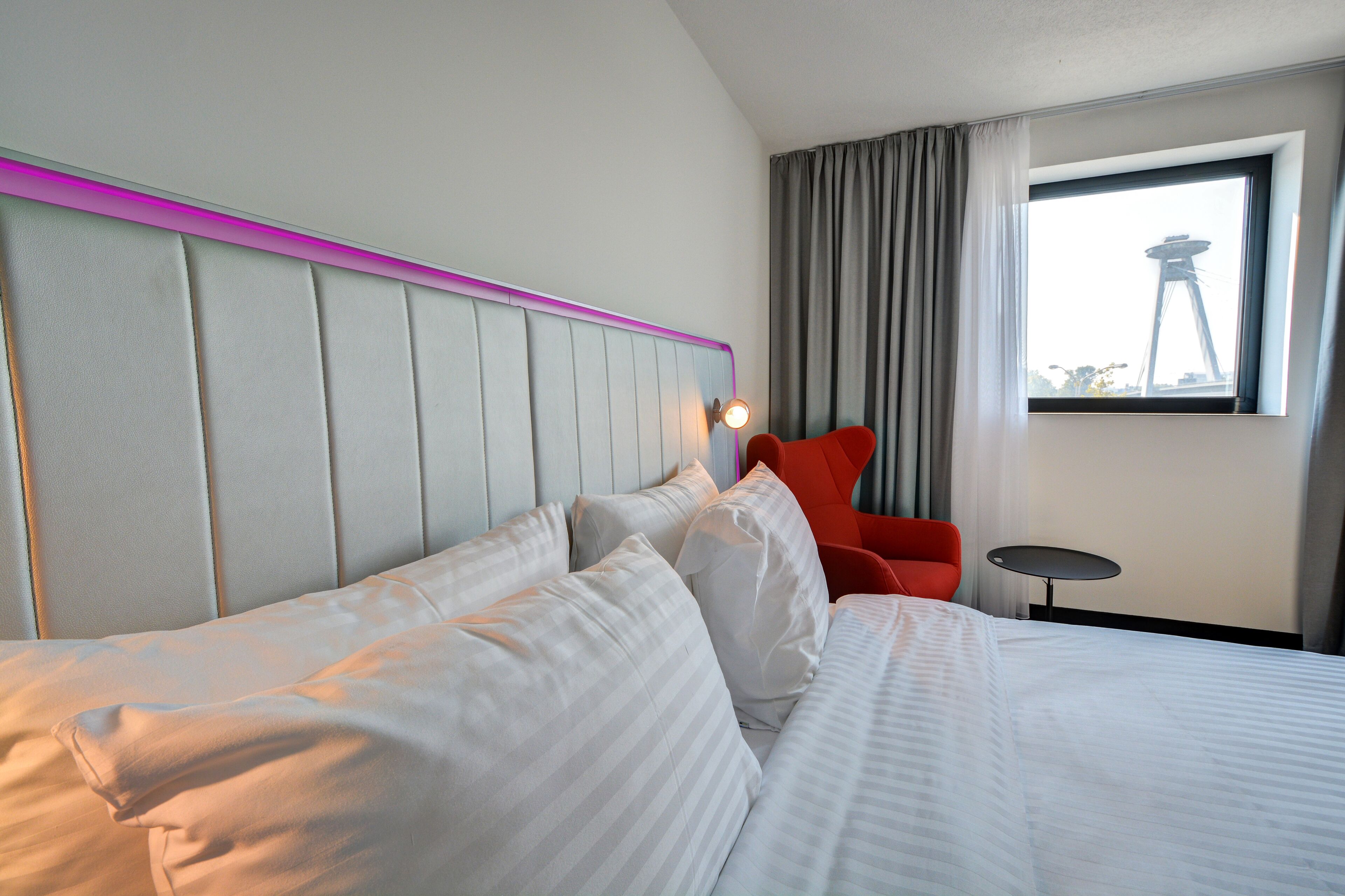 Una habitación de hotel con una cama grande, un cabecero con detalles rosados, una silla roja, una mesita pequeña y una ventana con vista a una torre.