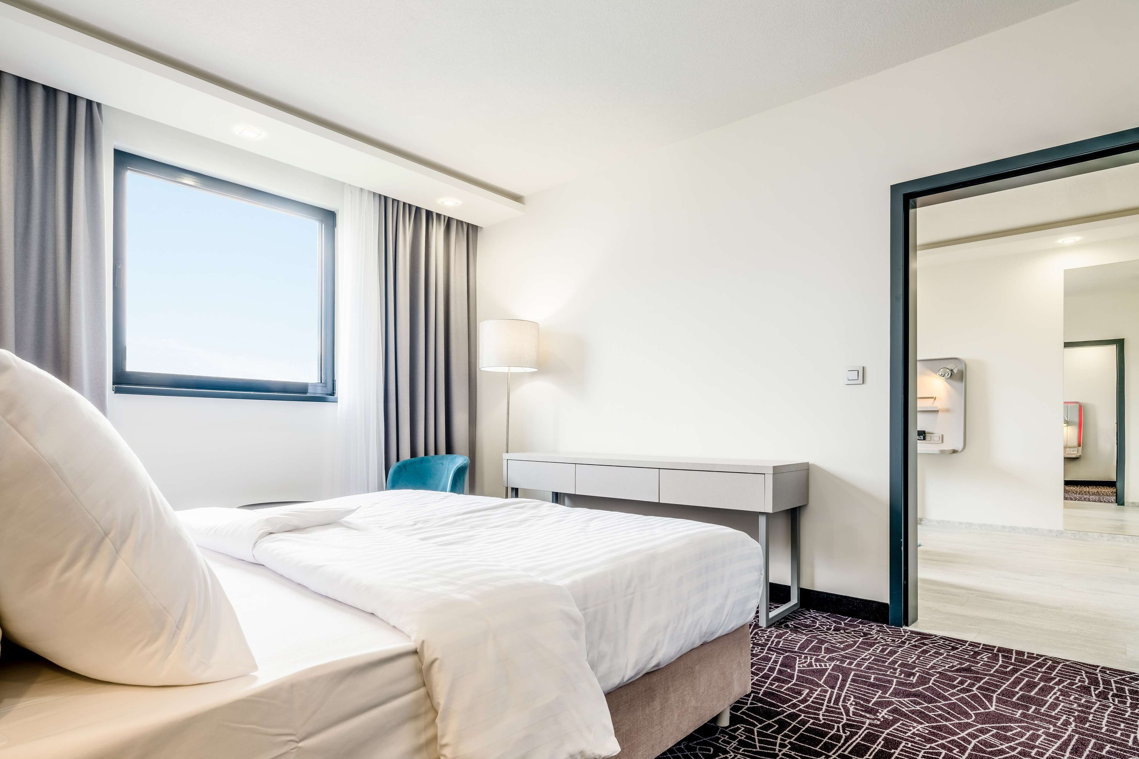 A hotel room with a bed, a desk, a chair, a lamp, and a view of the hallway.