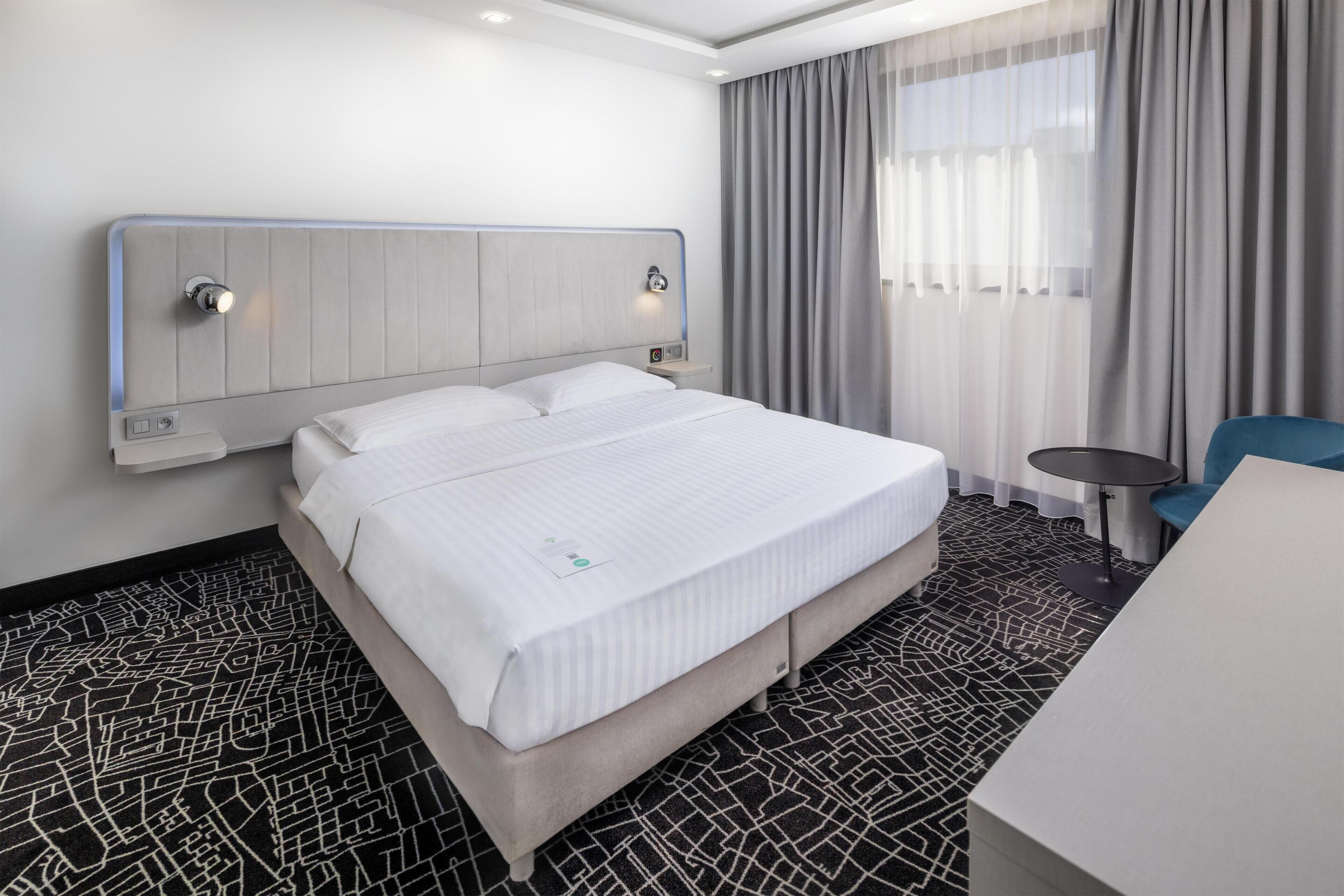 Een hotelkamer met een groot bed, een nachtkastje, een stoel, een bijzettafeltje en een raam met gordijnen.