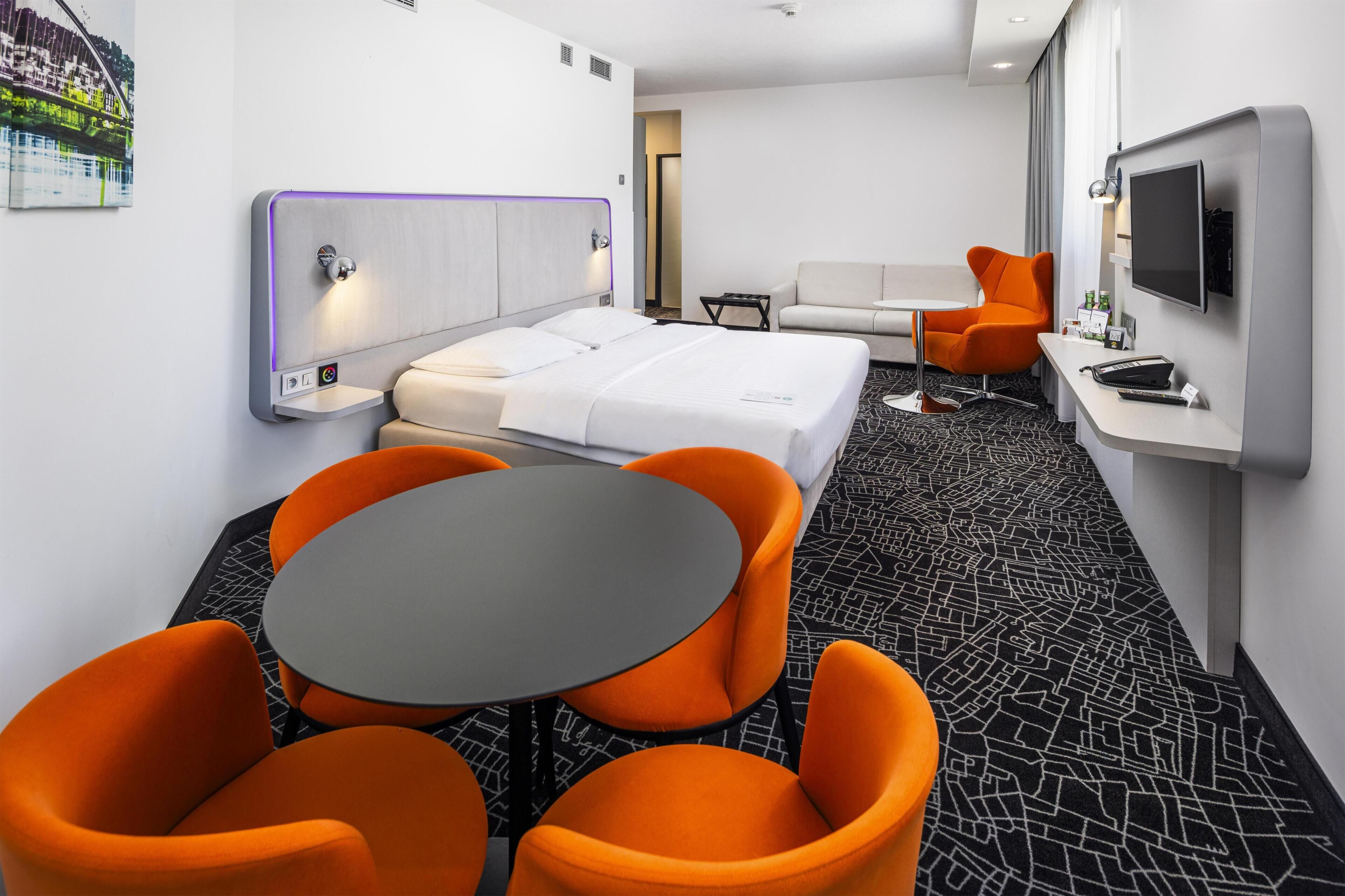 Una habitación de hotel con cama, escritorio, televisión y sillas naranjas.