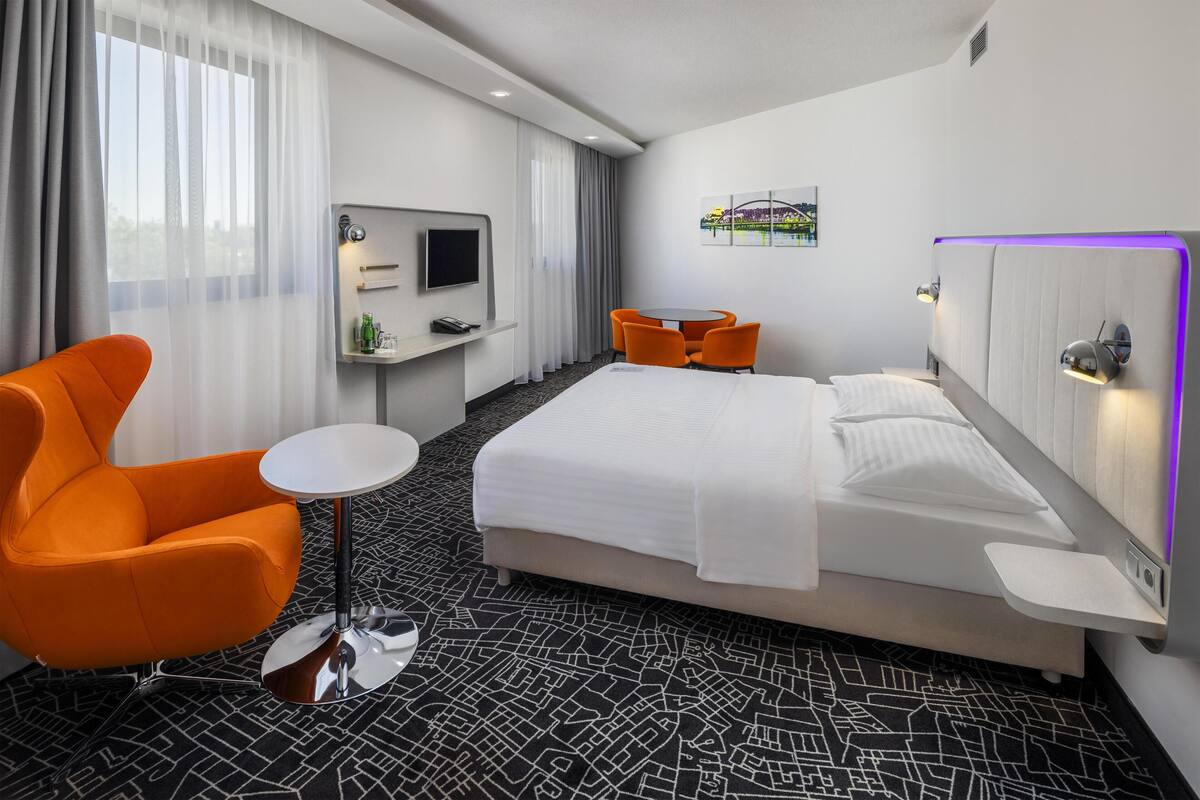 A hotel room with a bed, an orange chair, a small table, a TV, and a window with curtains.
