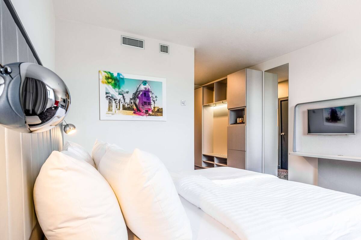 A hotel room with a bed, a large mirror, a picture on the wall, and a TV mounted on the wall.