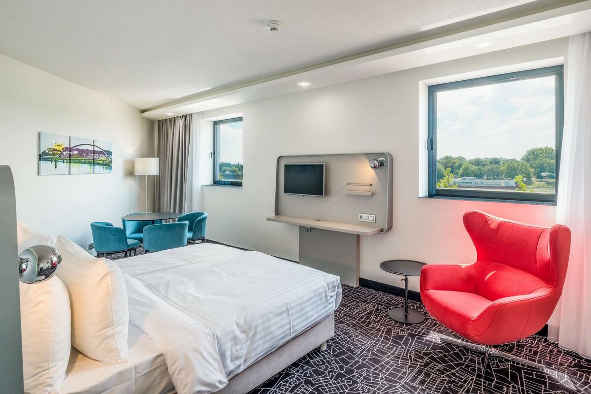 A hotel room with a bed, a TV, a red chair, a desk, and a view of the outdoors.