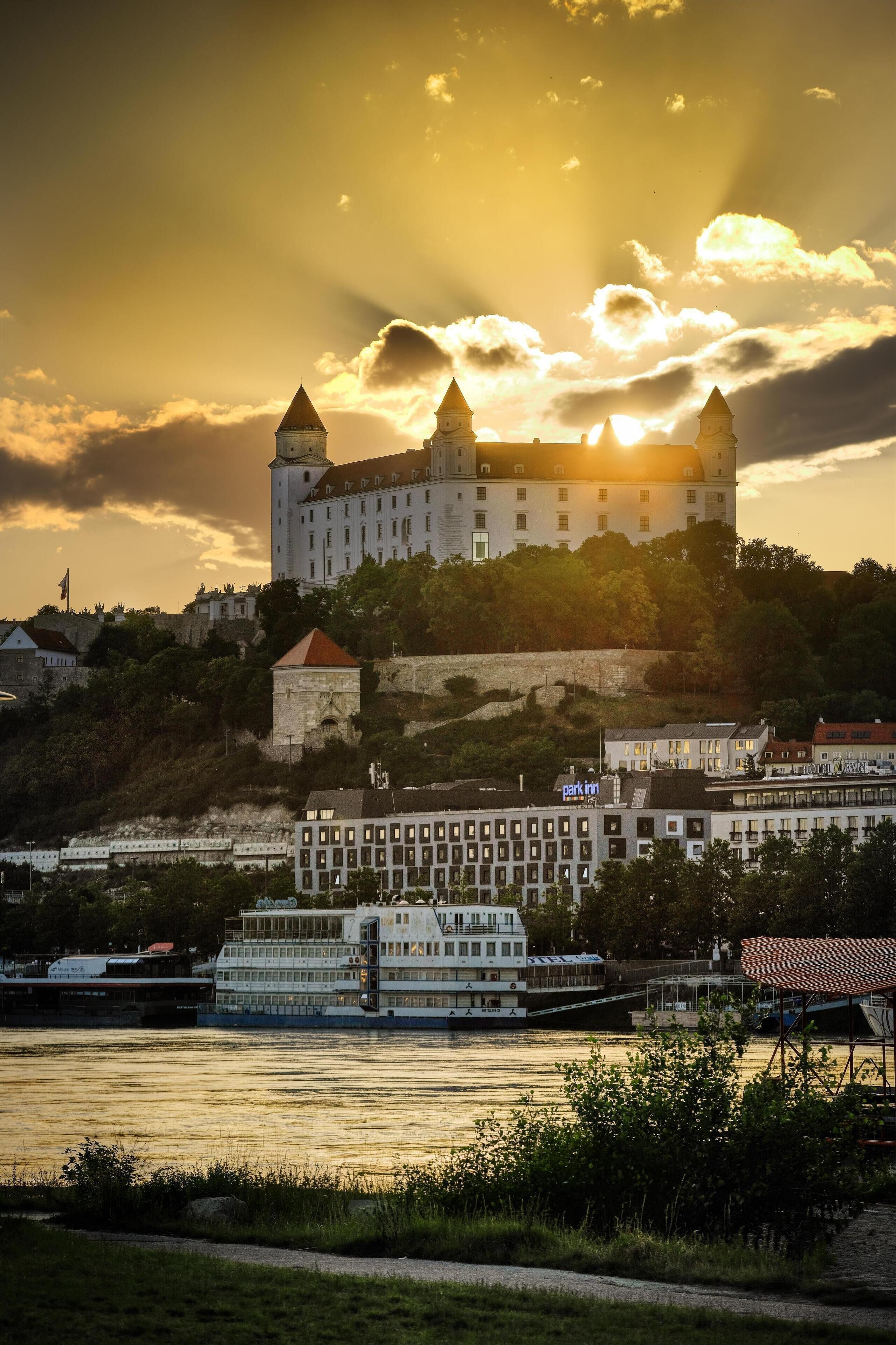 A historic castle with towers, a river, and a modern building with a sign.