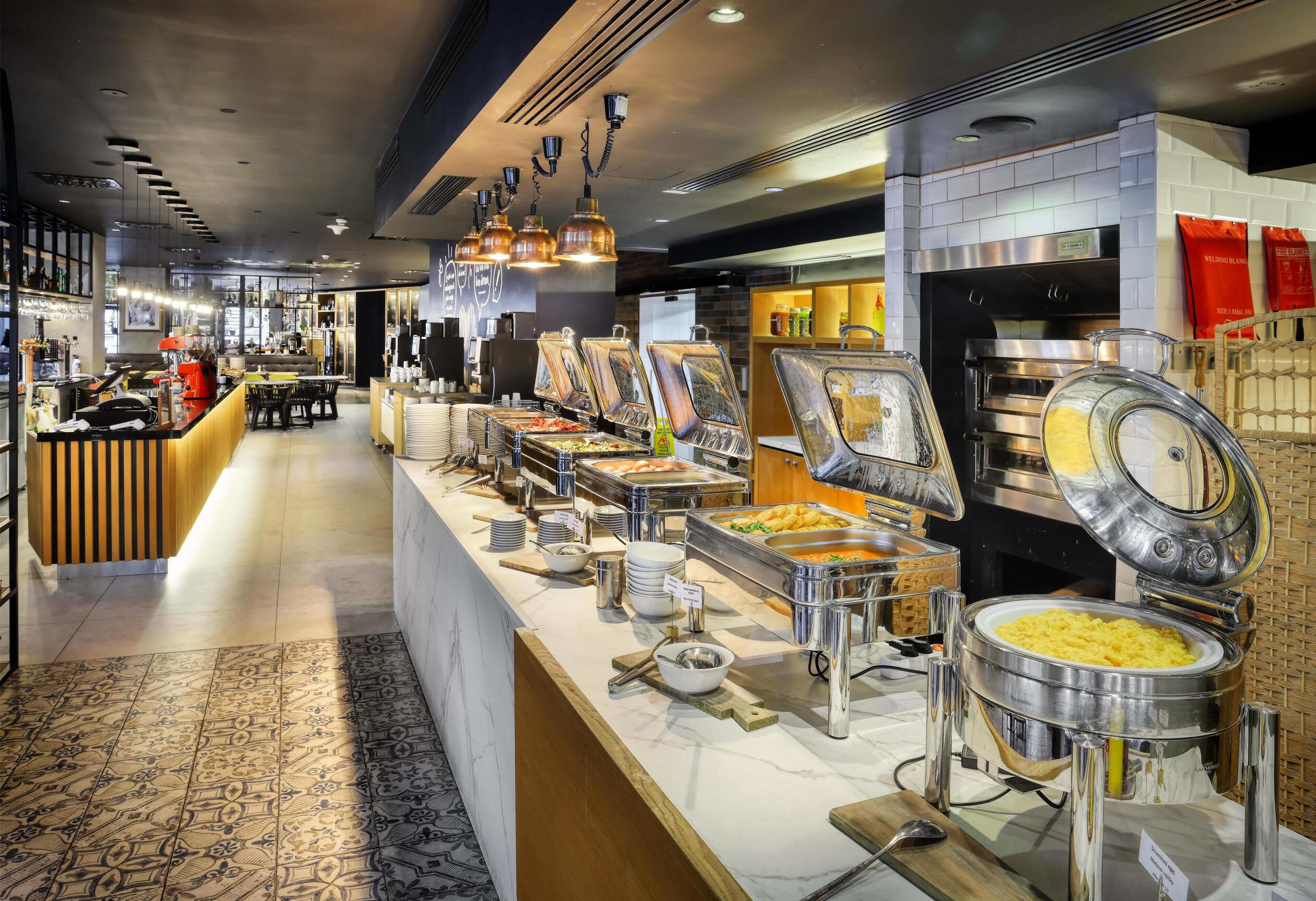 Uma área de buffet com diversos pontos de comida, um espaço para sentar e um balcão com máquina de café.