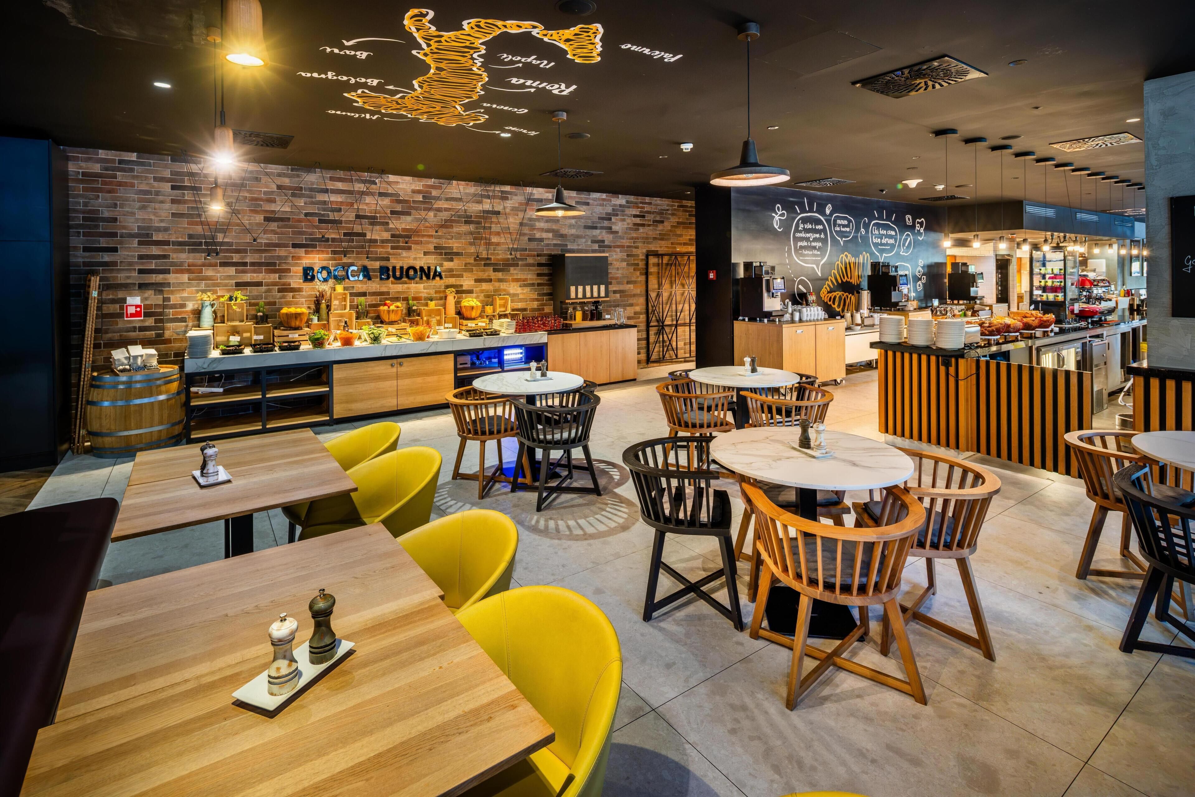 A restaurant interior with a brick wall, wooden tables, yellow chairs, and a bar area.