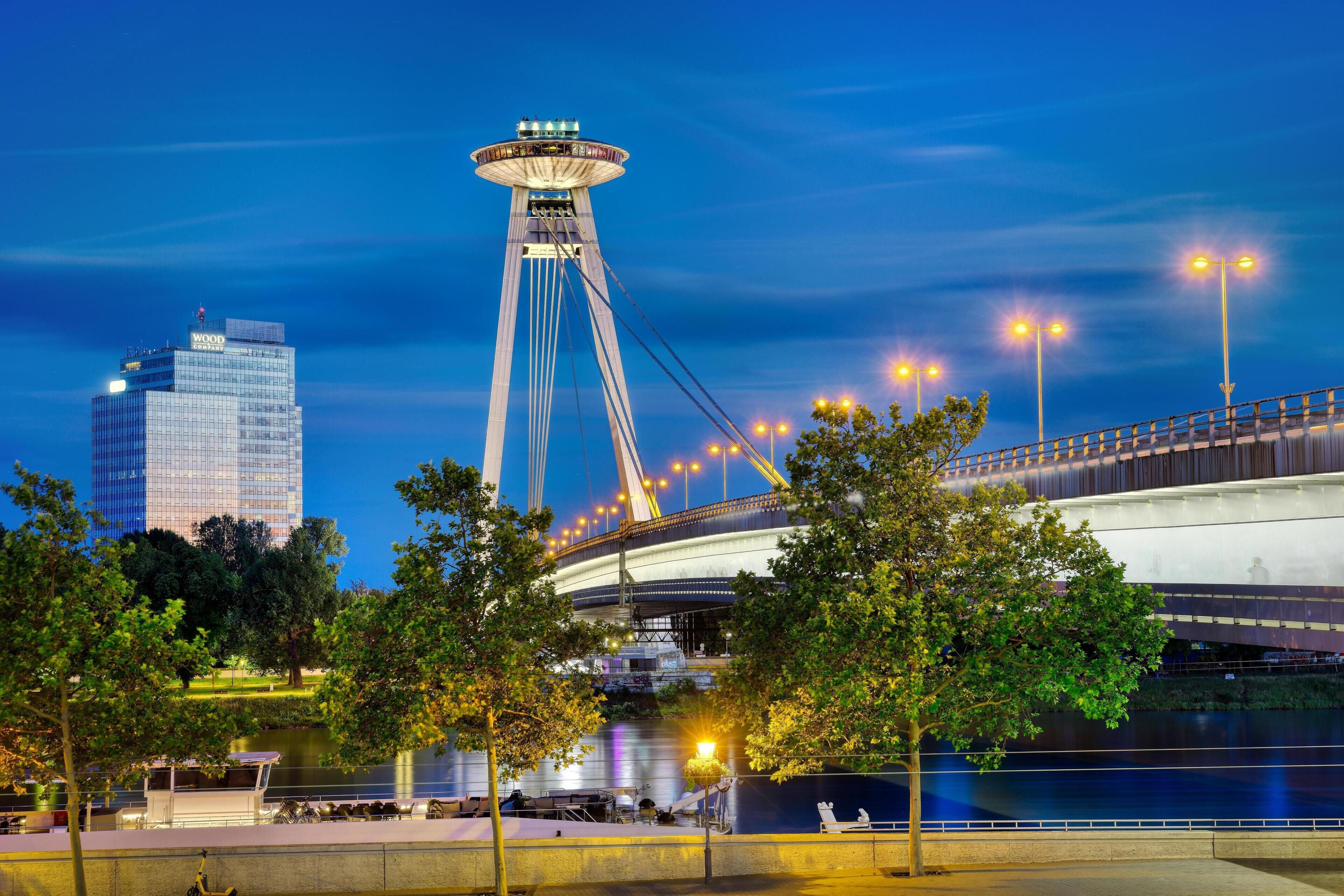 A nighttime view of a city with a prominent cable-stayed bridge, a modern building with a glass facade, and a waterfront area with seating.