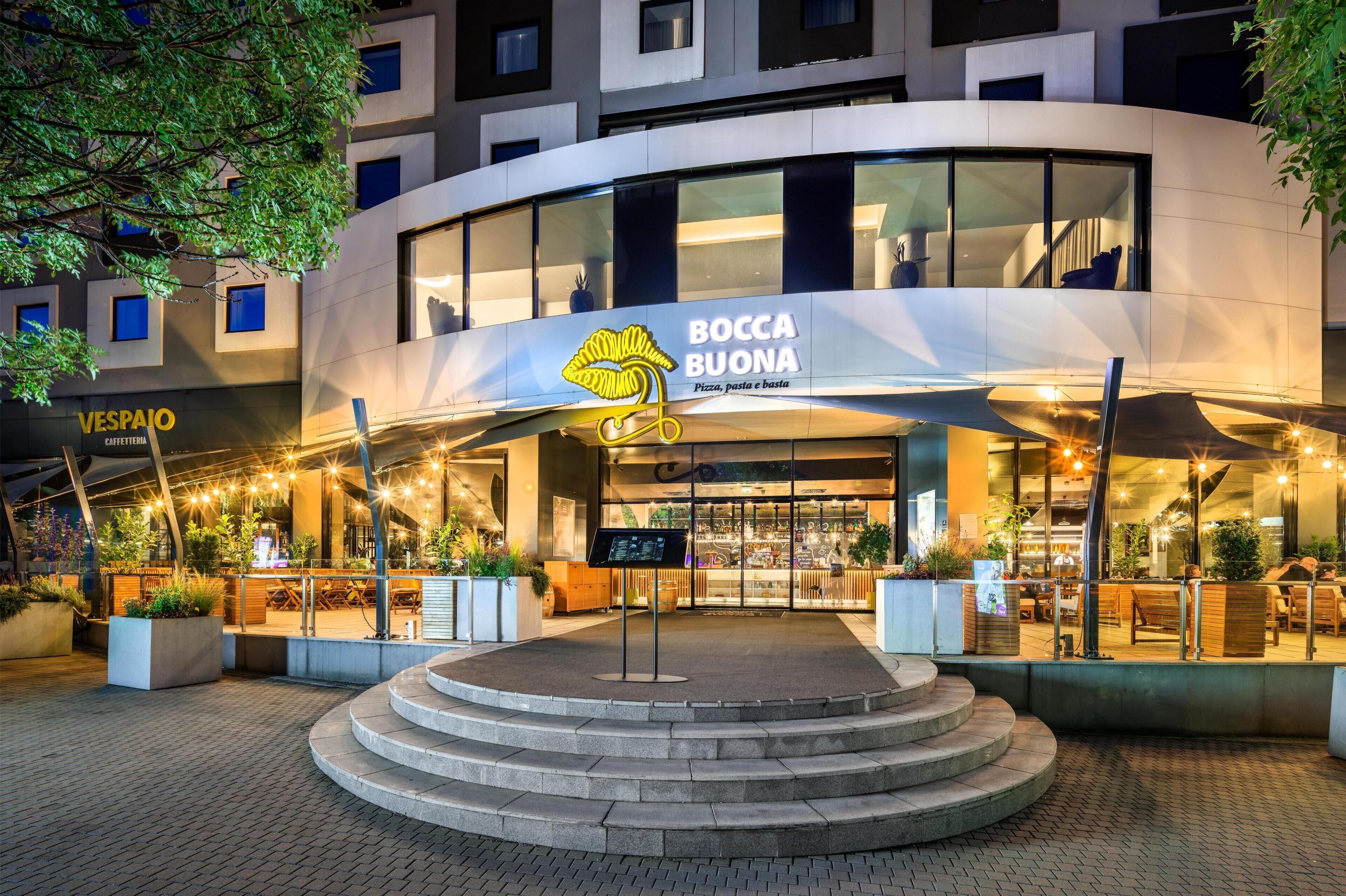 A modern building with a restaurant named 'BOCCA BUONA' featuring a pizza, pasta, and bread sign.