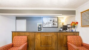 Un mostrador de recepción con un cartel de bienvenida enmarcado, dos sillones estampados, una mesita con un jarrón de flores y un reloj fijado a la pared.