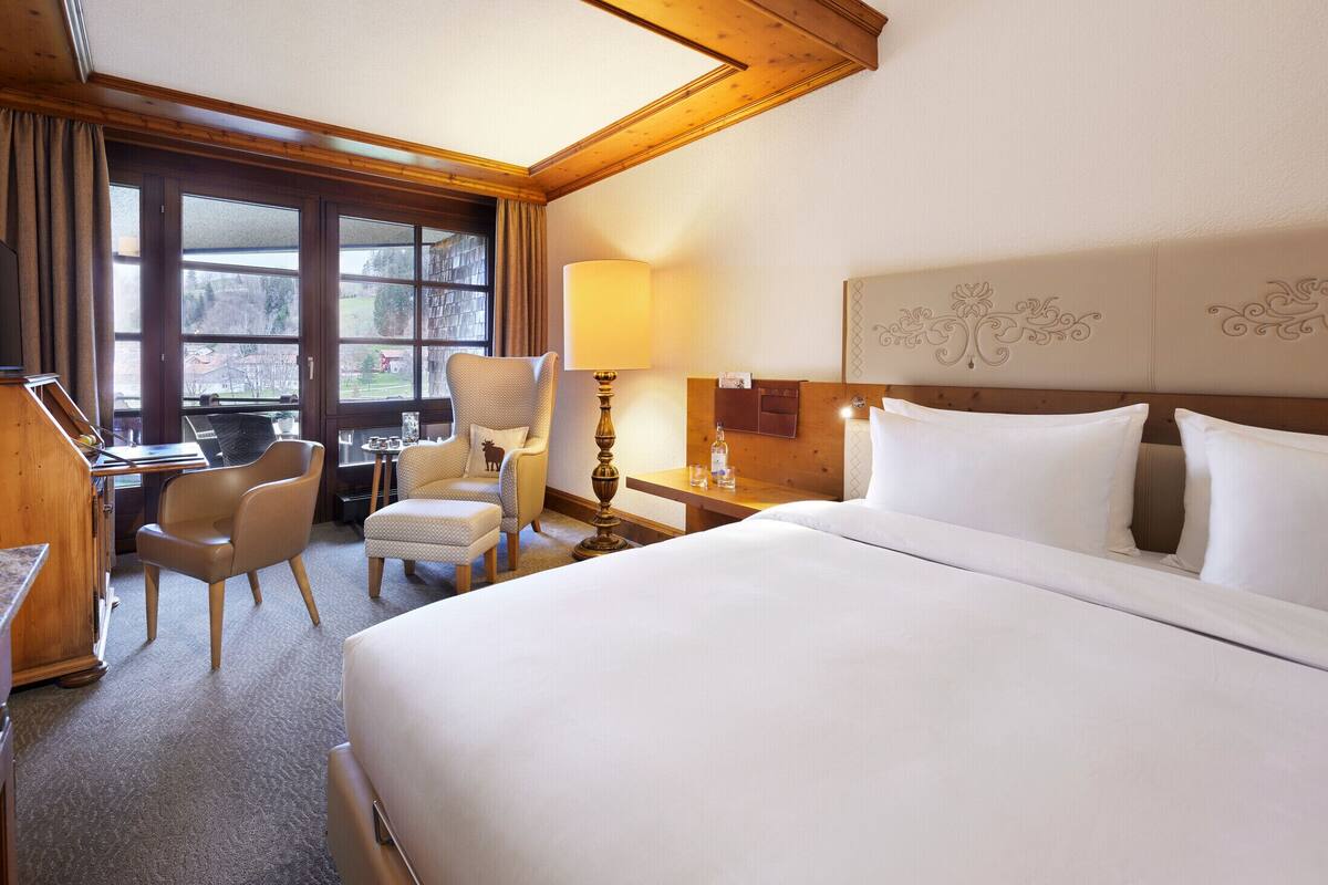 A hotel room with a large bed, a desk, two chairs, a lamp, and a window with curtains.