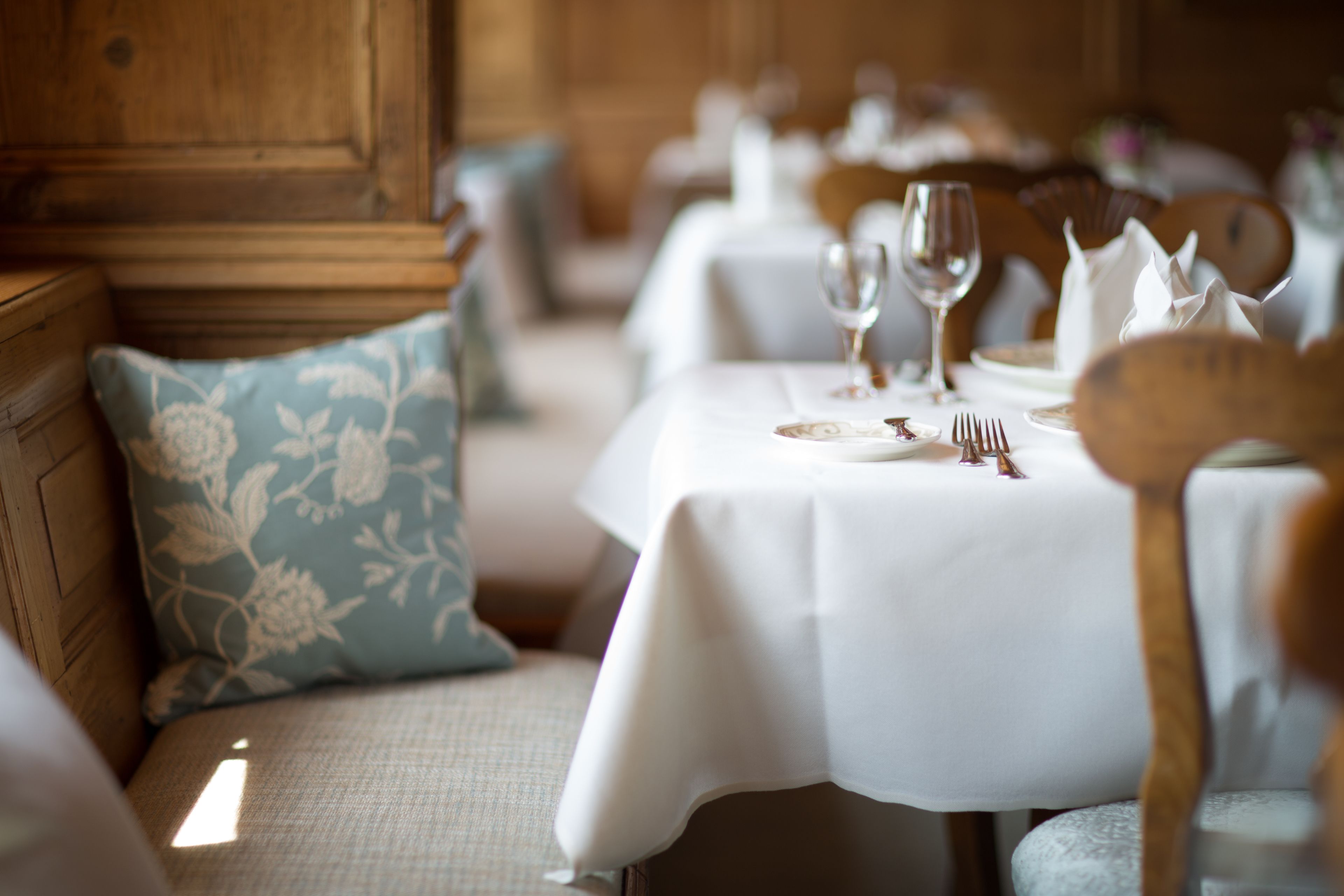 A dining area with tables set for a meal, featuring white tablecloths, wooden chairs, and floral cushions.