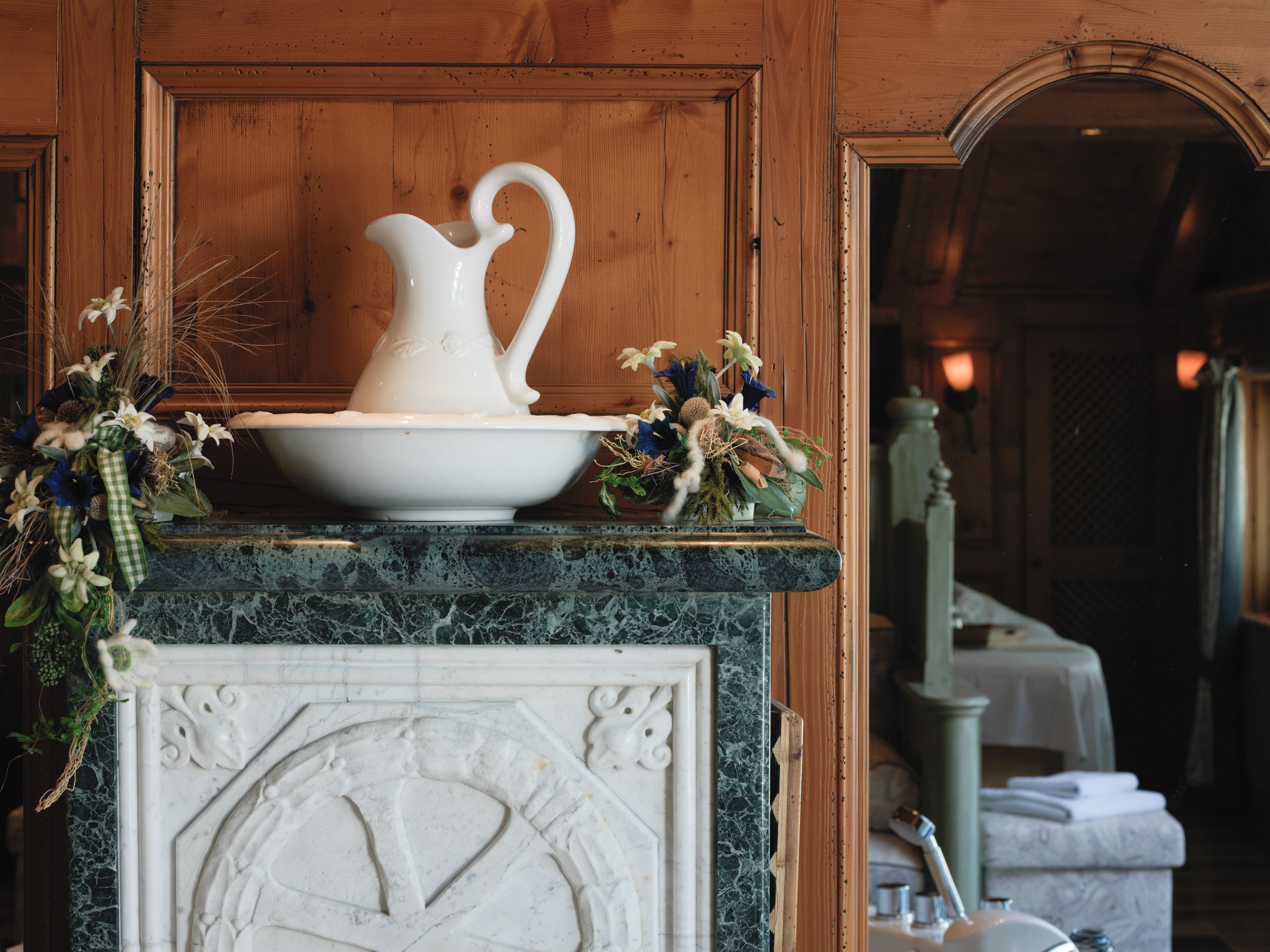 A marble fireplace with a pitcher and bowl, surrounded by floral arrangements.