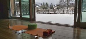 Enjoy a serene view of the surrounding landscape from the large window, featuring a church and a fence. The room is equipped with a wooden floor, a cushioned stool, a yoga mat, and a candle.
