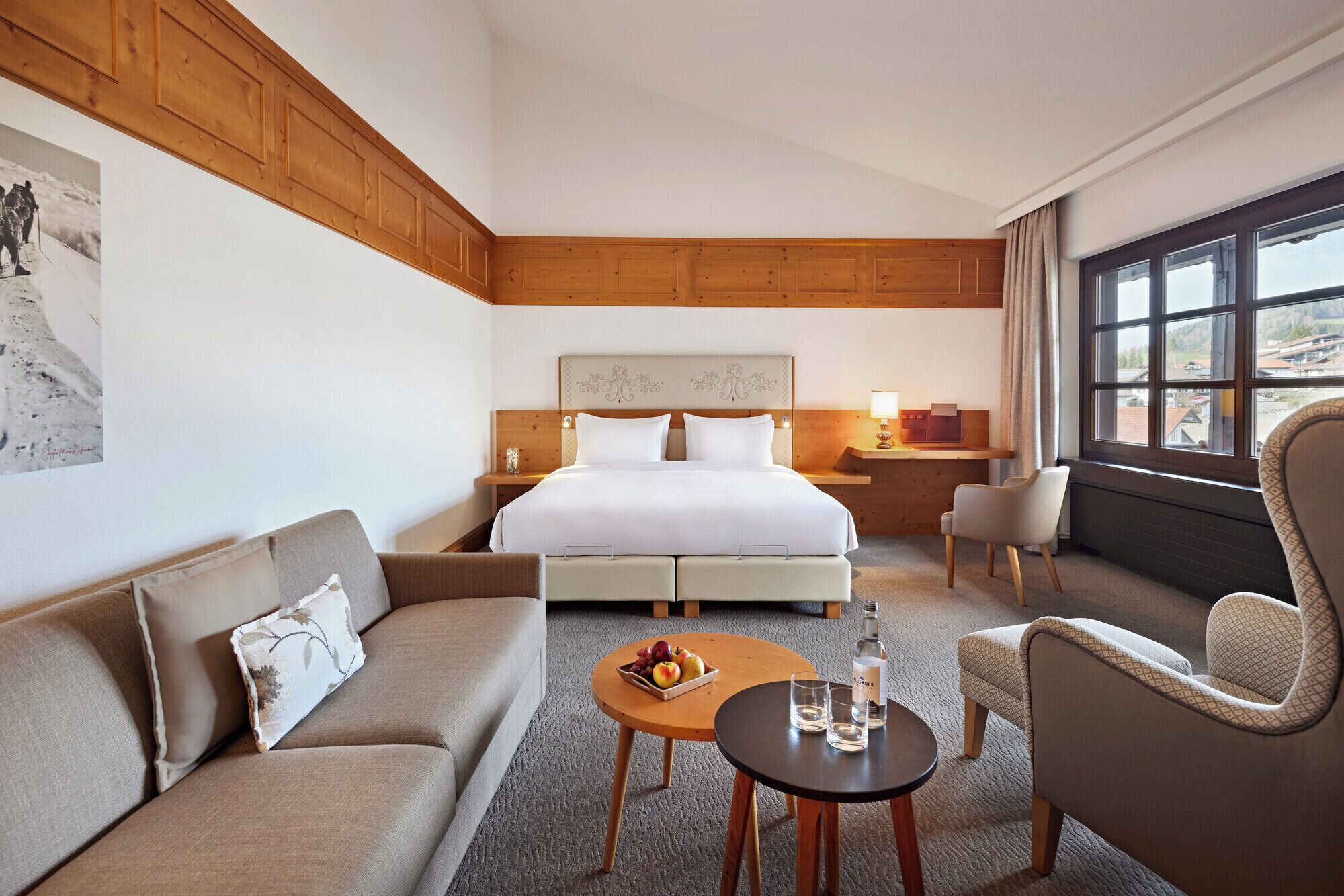 Habitación de hotel con cama, sofá, sillón, una mesita con fruta y vasos, y ventana con vistas.