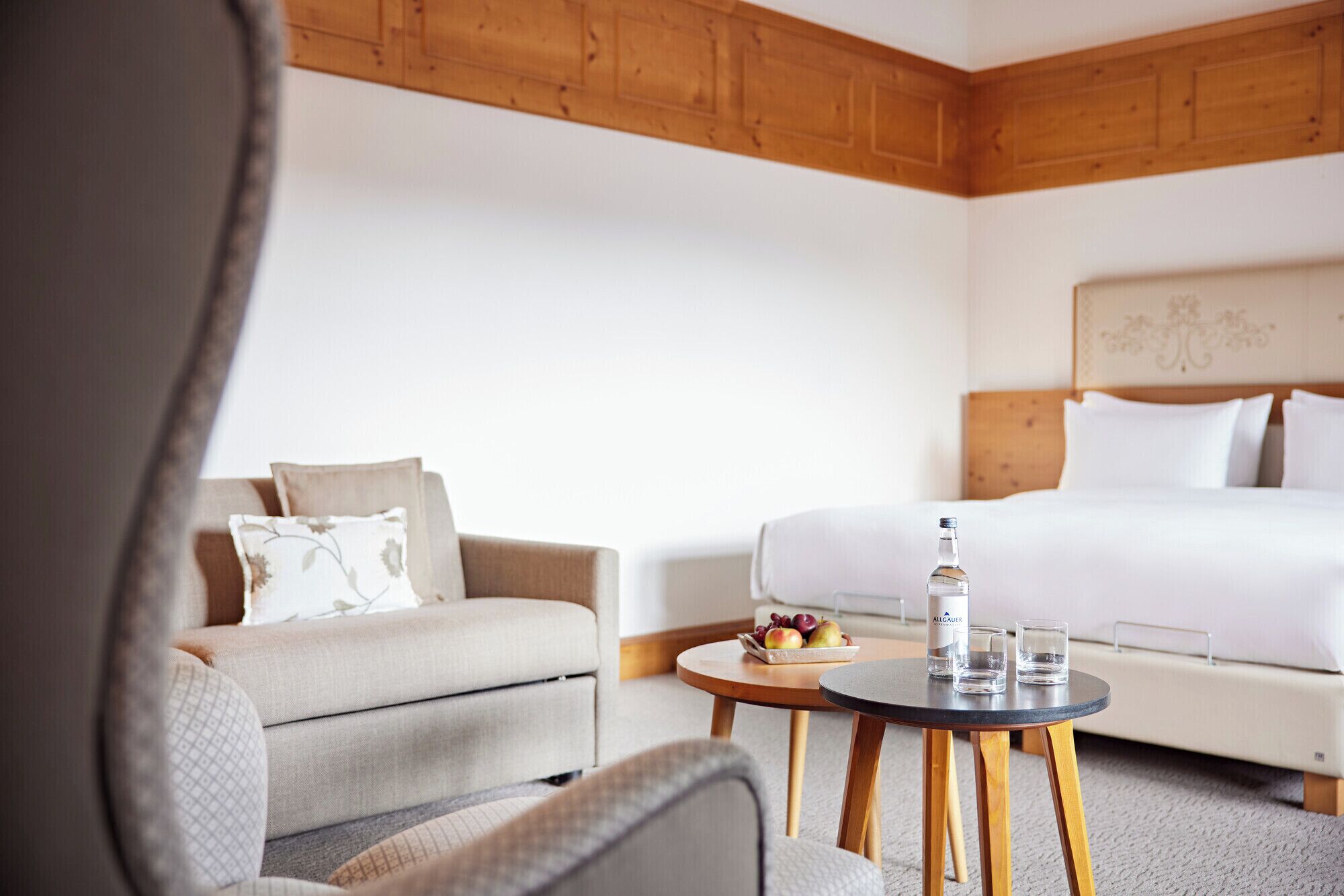 Una habitación de hotel con cama, sofá, una mesita redonda con una botella y vasos, y un frutero con fruta.