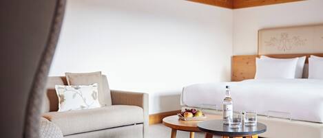 Una habitación de hotel con cama, sofá, una mesita redonda con una botella y vasos, y un frutero con fruta.