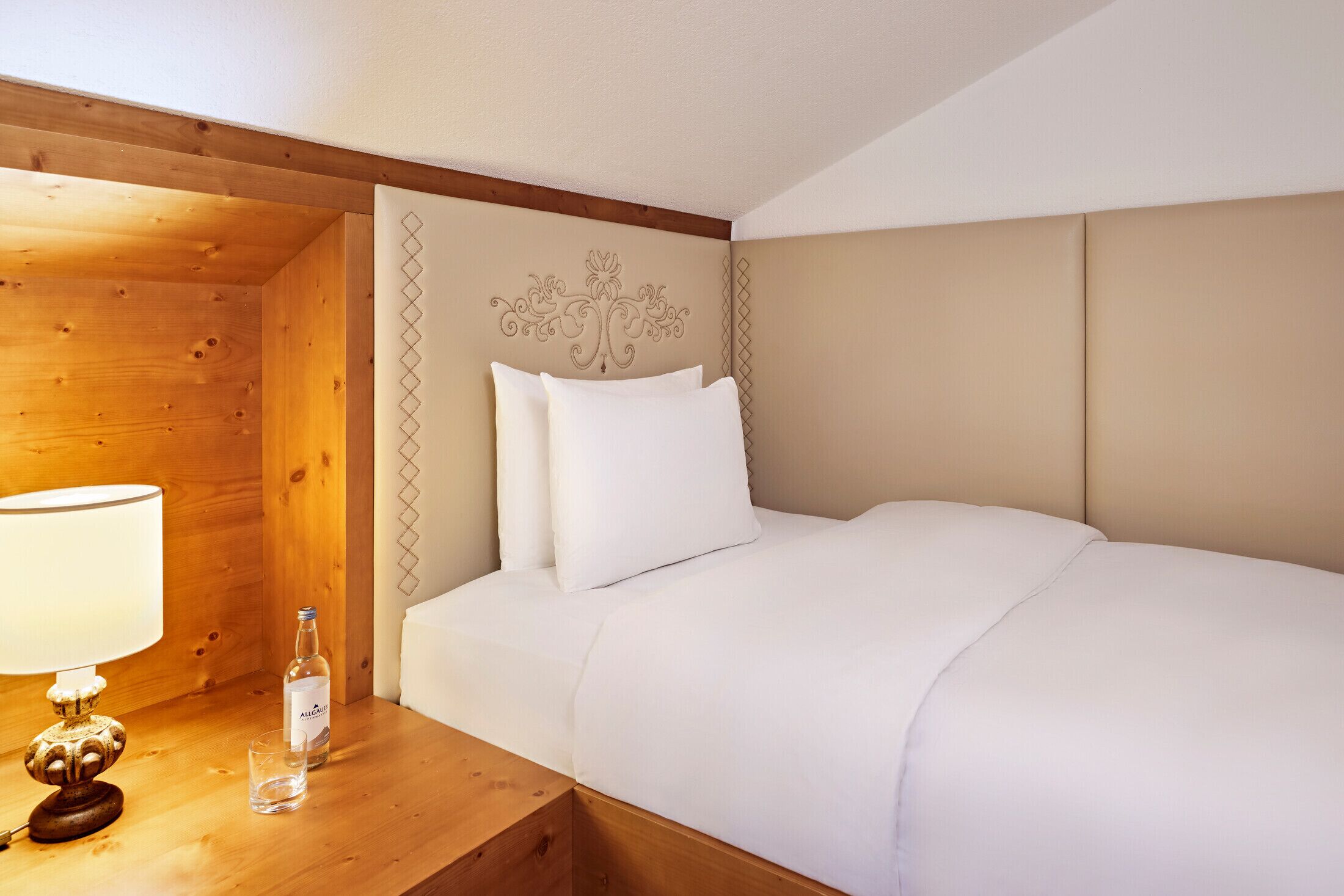 Un lit bien fait, avec du linge de lit blanc, une table de chevet en bois sur laquelle se trouvent une lampe et une bouteille, et une tête de lit décorative.