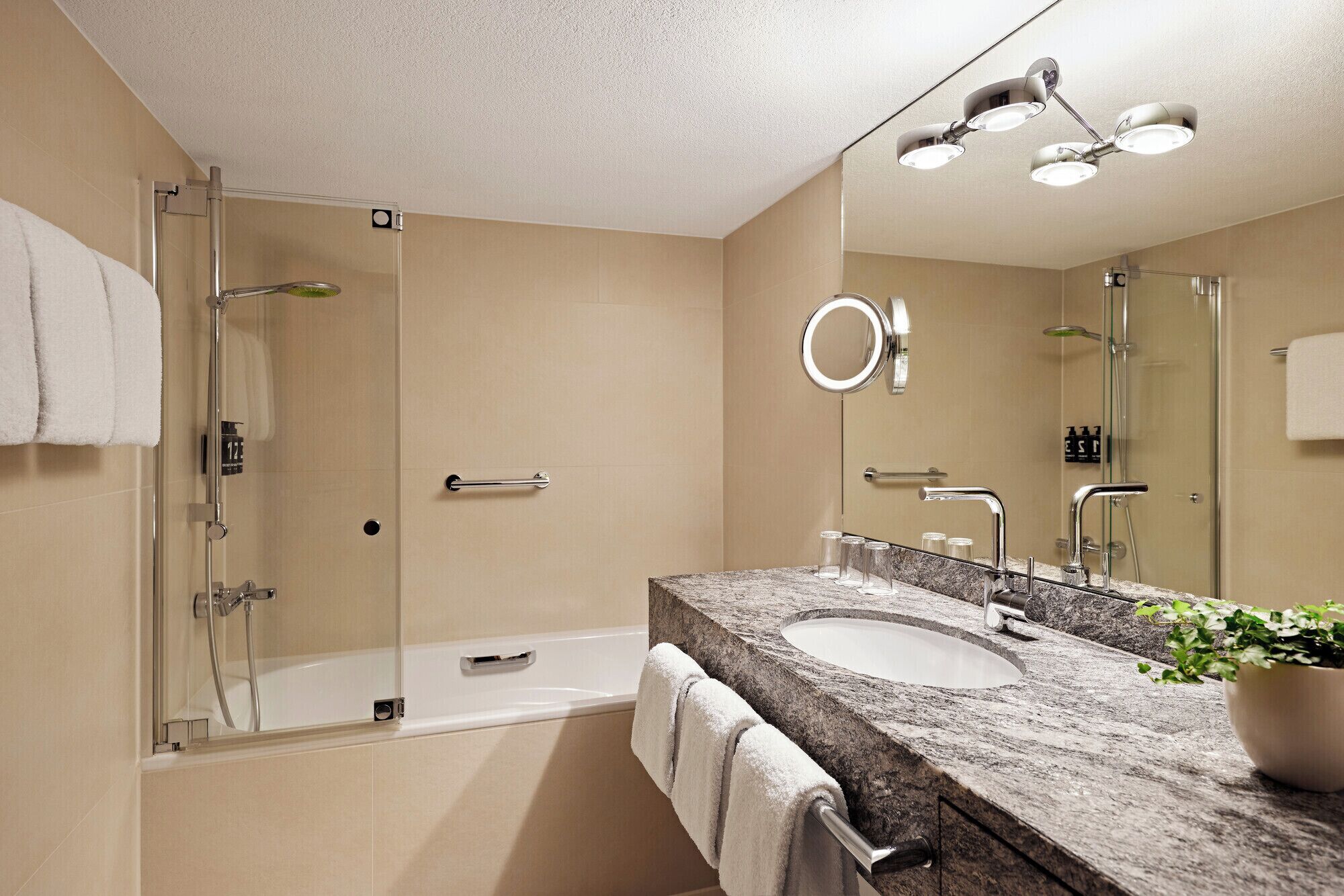 A modern bathroom with a glass shower enclosure, a bathtub, a granite countertop sink, and a large mirror with a circular light.